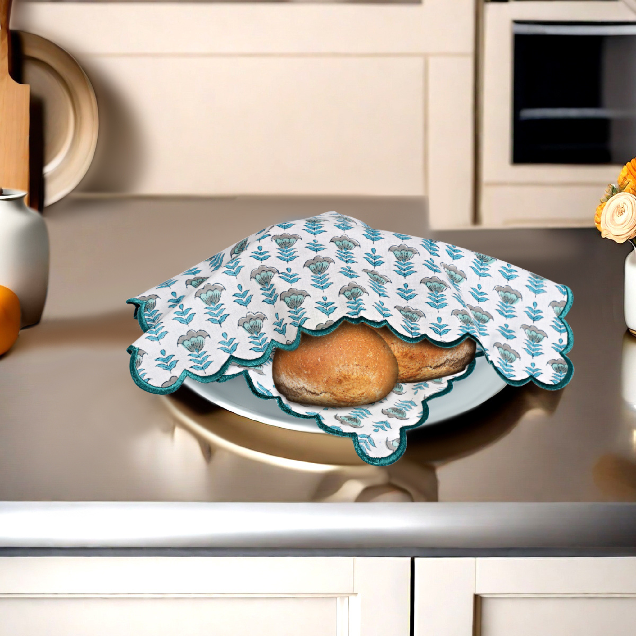 Decorative cloth with floral pattern covering a plate of bread on a kitchen counter.