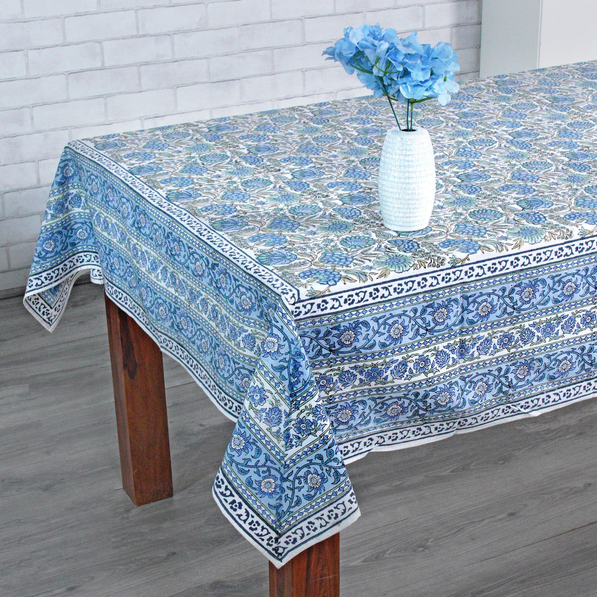 Tablecloth with blue floral pattern on a wooden table against a white brick wall.