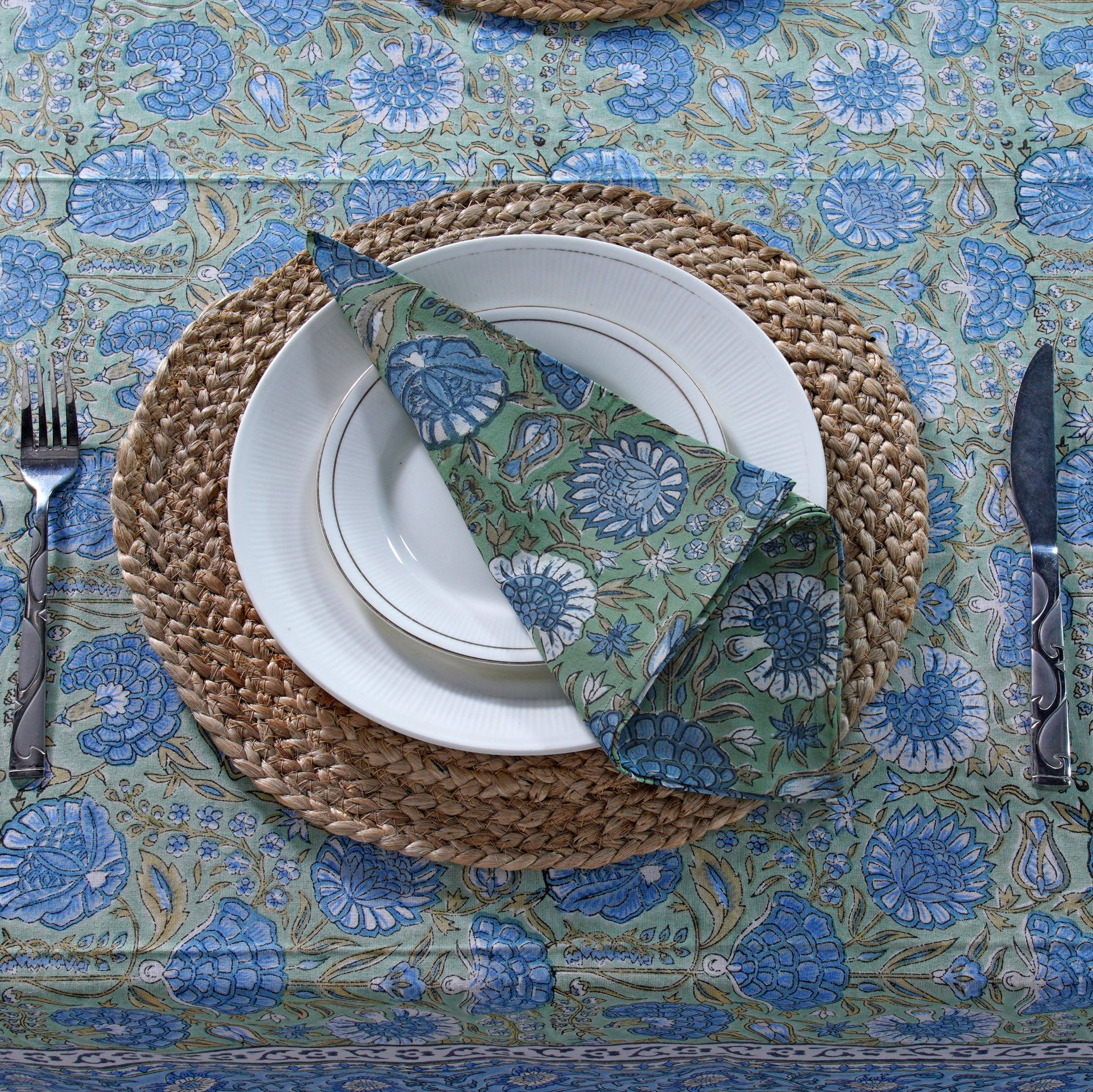 Dining table setting with a blue and green patterned tablecloth, white plates, and cutlery.