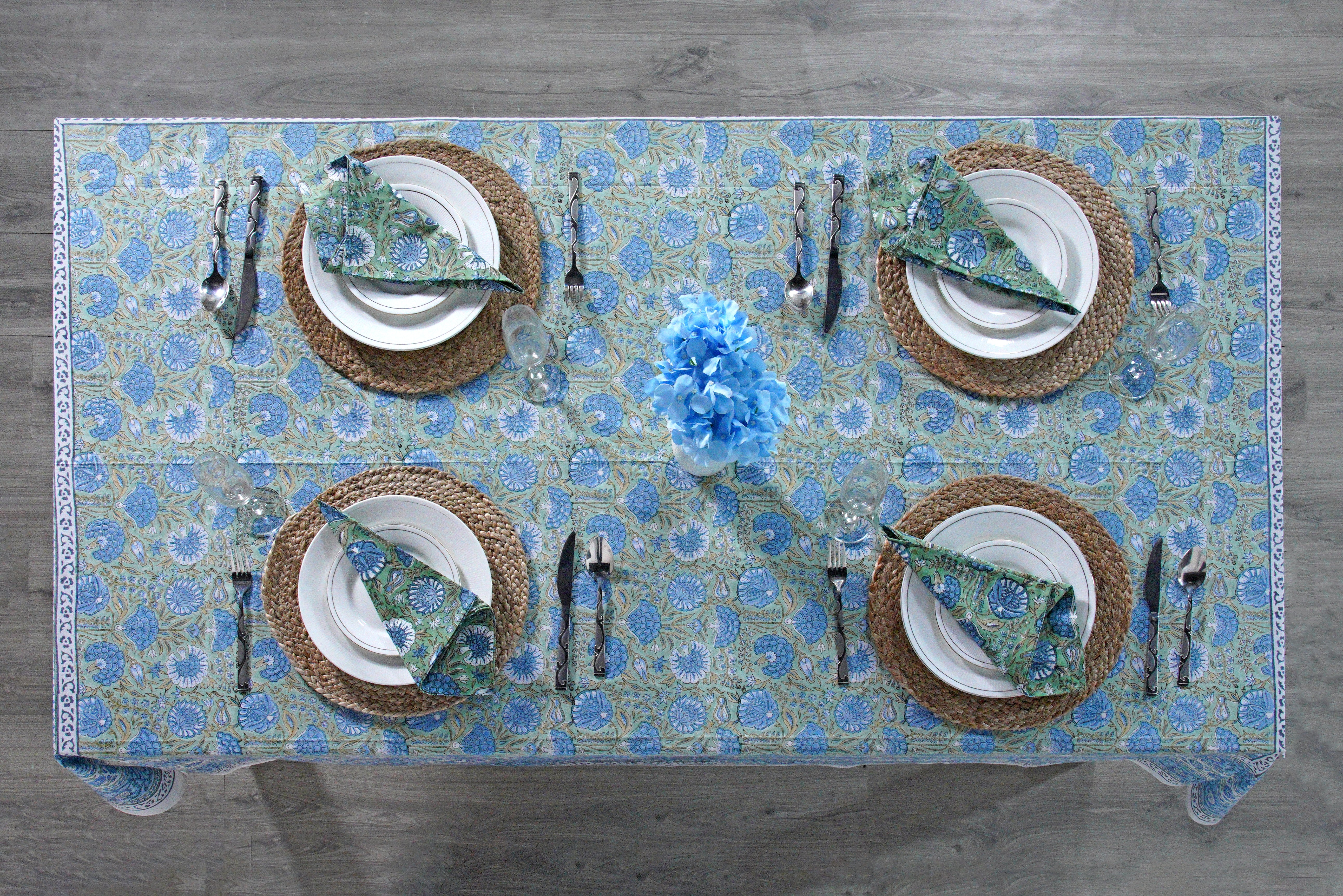 Table setting with floral tablecloth, plates, cutlery, and napkins on a wooden surface.
