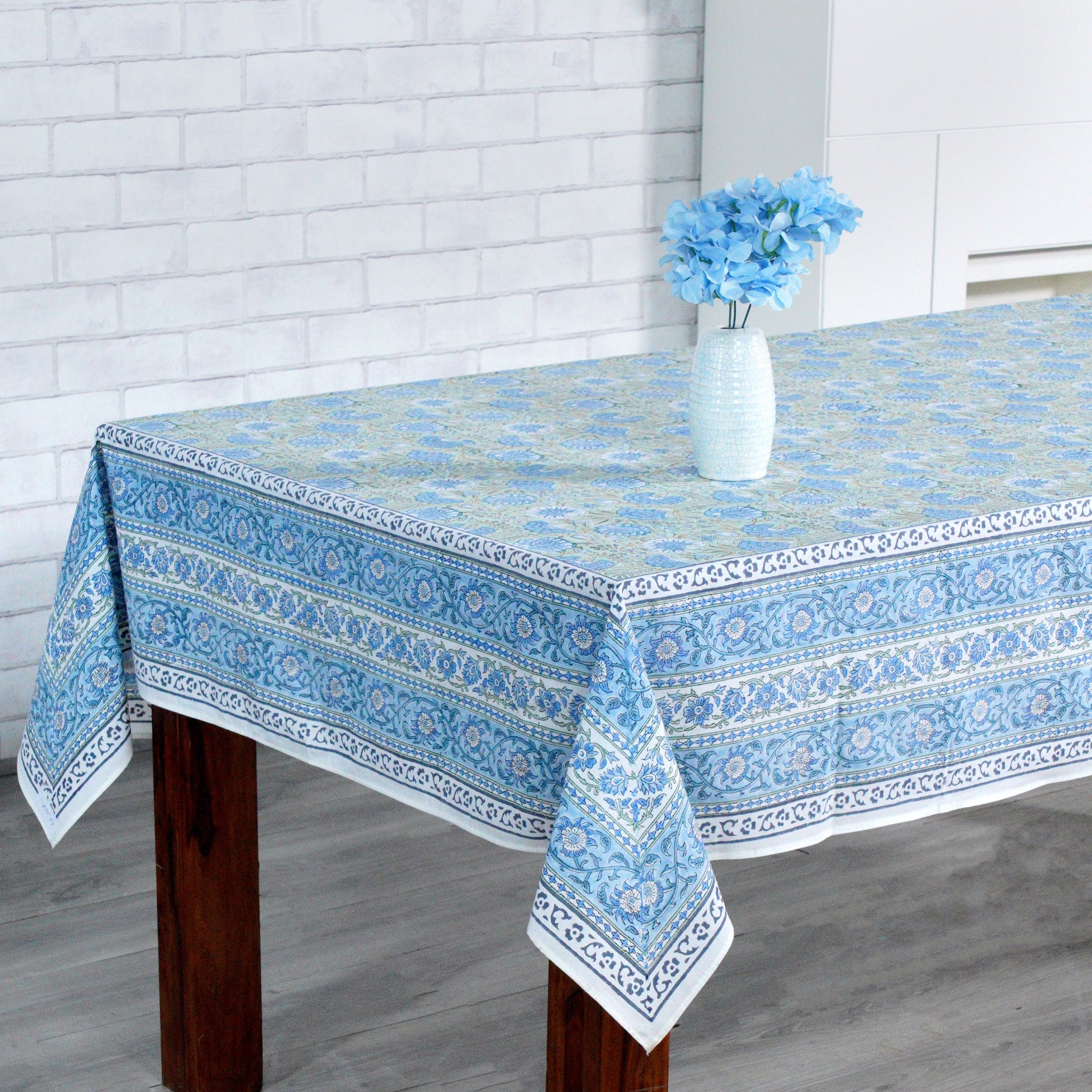 Tablecloth with blue floral pattern on a wooden table against a white brick wall.