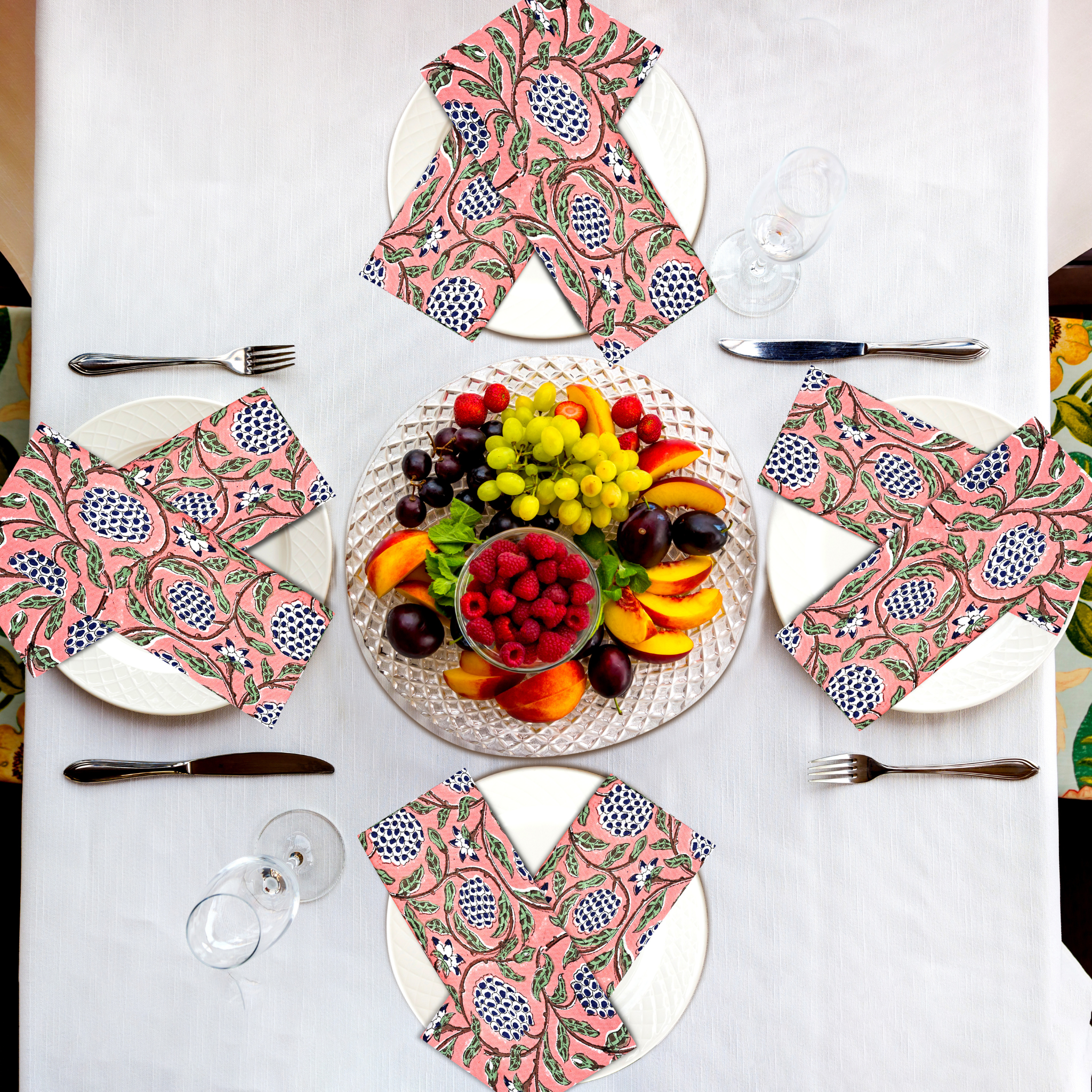Table setting with pink patterned napkins, fruit platter, and cutlery on a white tablecloth.