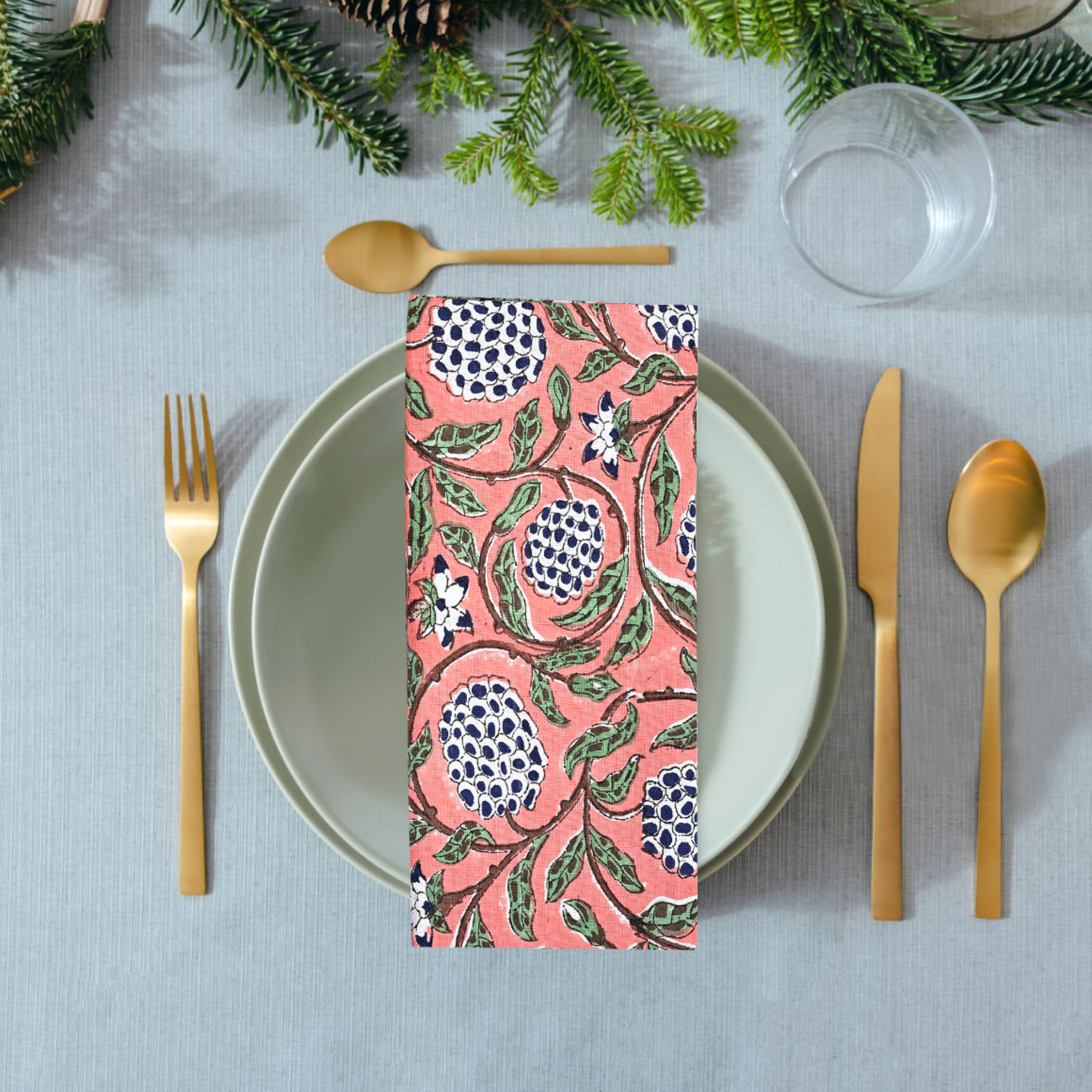 Table setting with a pink floral napkin, gold cutlery, and greenery on a gray tablecloth.