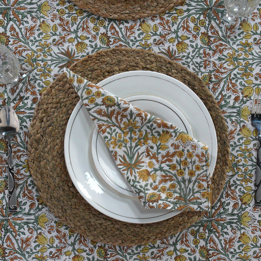 Table setting with floral napkin, white plates, and woven placemat on a patterned tablecloth.