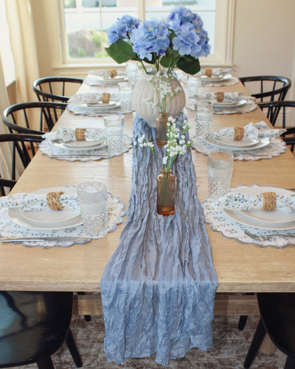 Dining table set with a blue lace runner, white plates, and a vase of blue flowers.