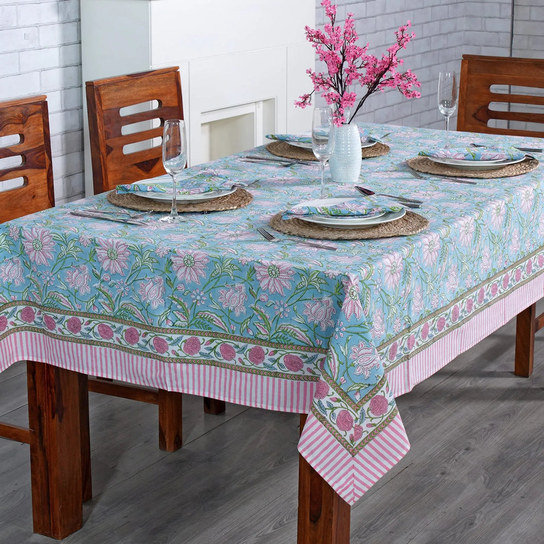 Dining table set with a floral tablecloth in a room with wooden chairs and a fireplace.