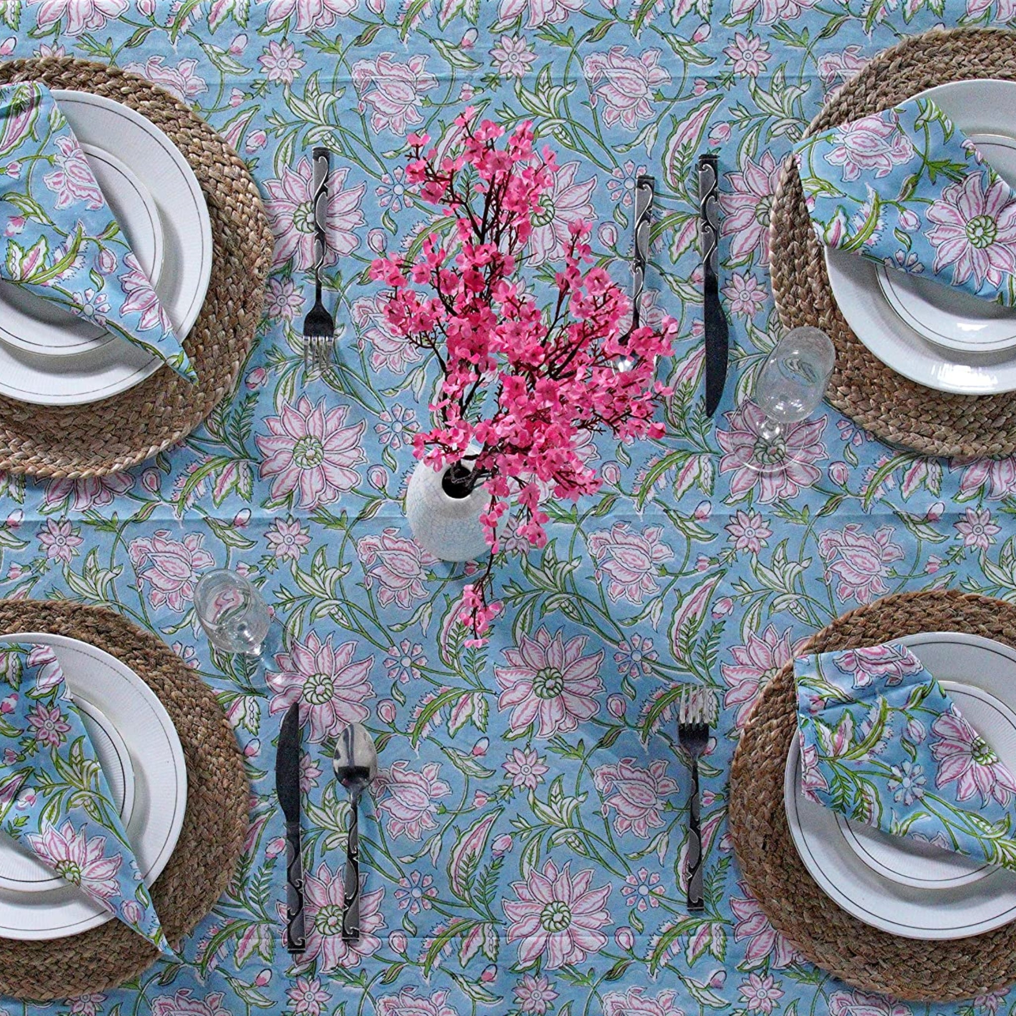 Table setting with floral tablecloth, plates, cutlery, and a vase of pink flowers.