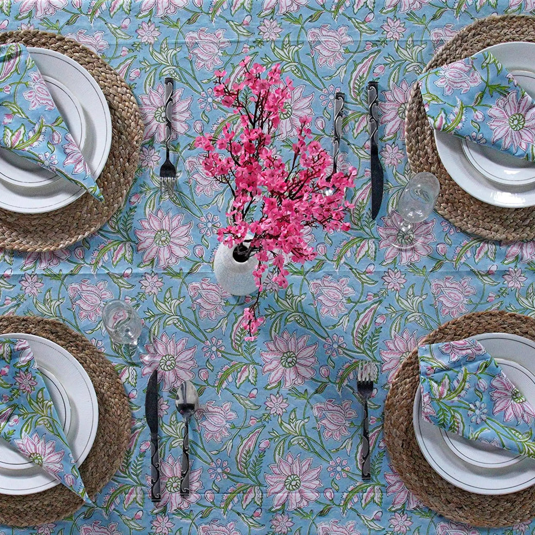Table setting with floral tablecloth, plates, cutlery, and a vase of pink flowers.