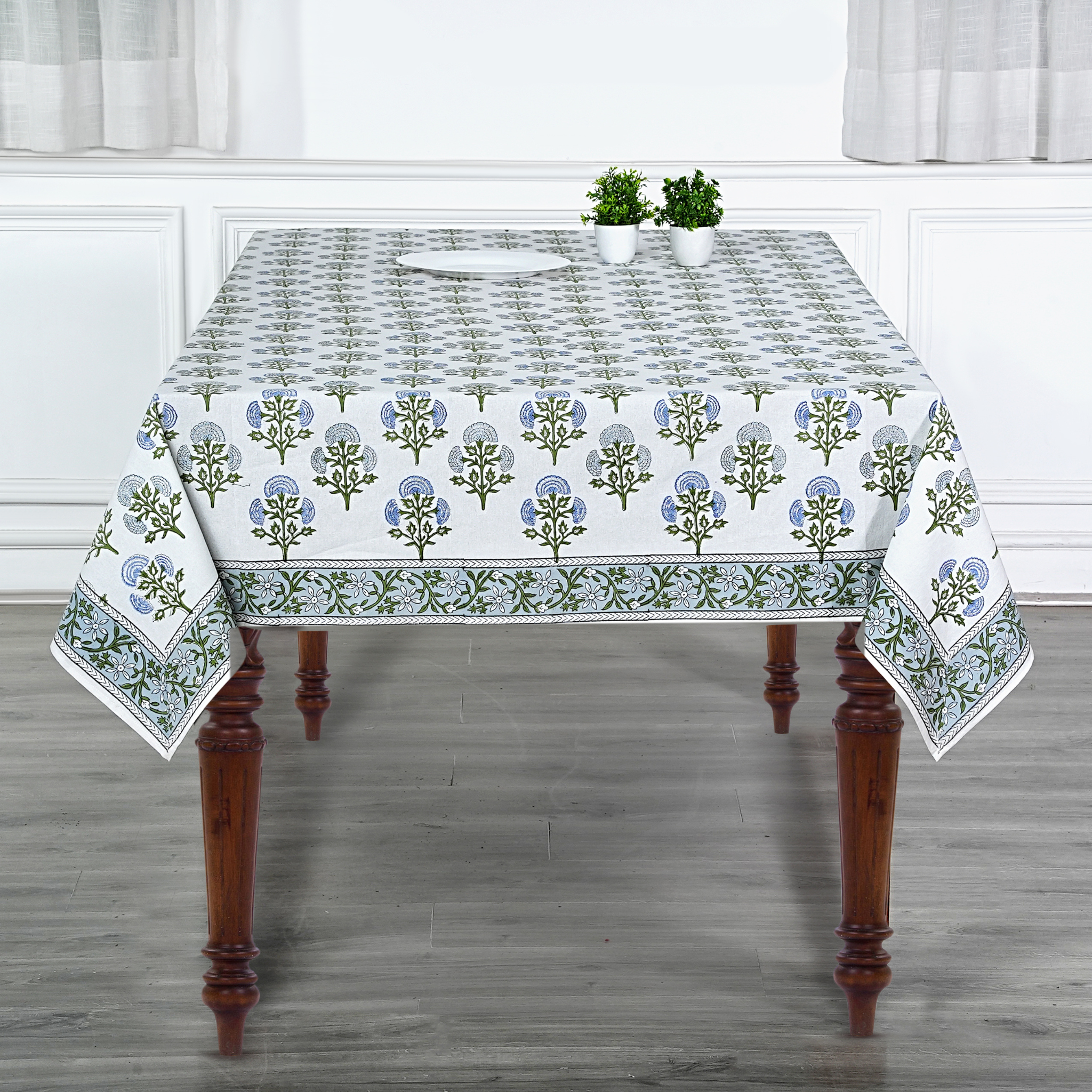 Tablecloth with floral pattern on a wooden table in a room with white walls and curtains.