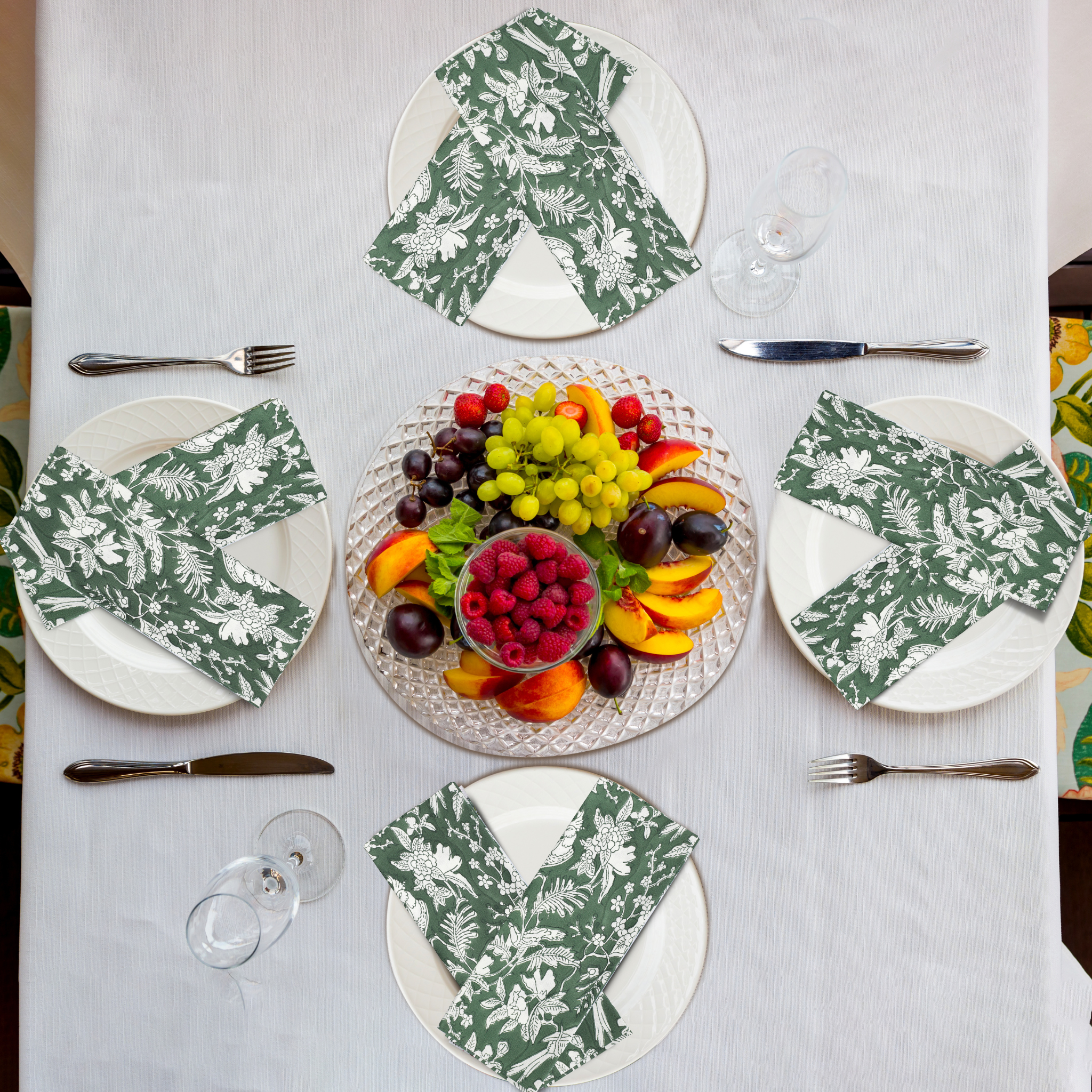 Table setting with floral napkins, fruit platter, and cutlery on a white tablecloth.