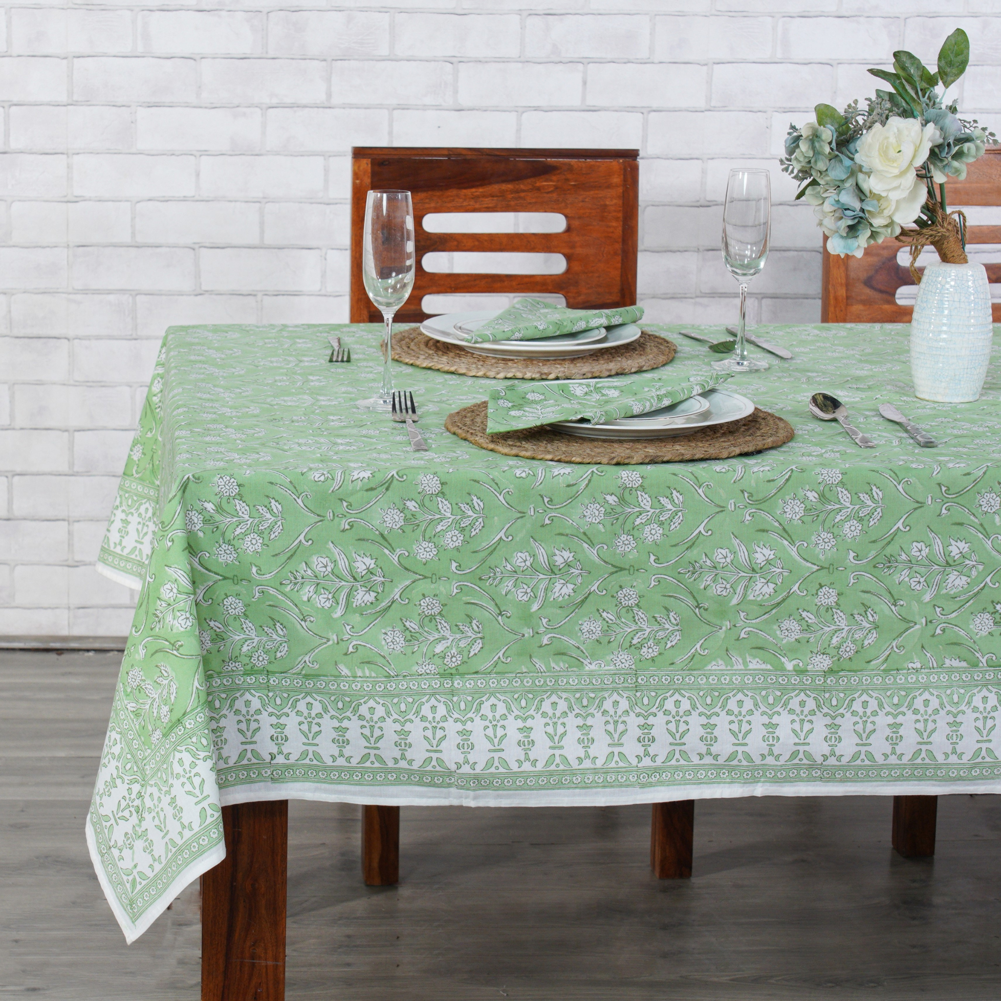 Dining table set with a green and white patterned tablecloth, wooden chairs, and white plates.