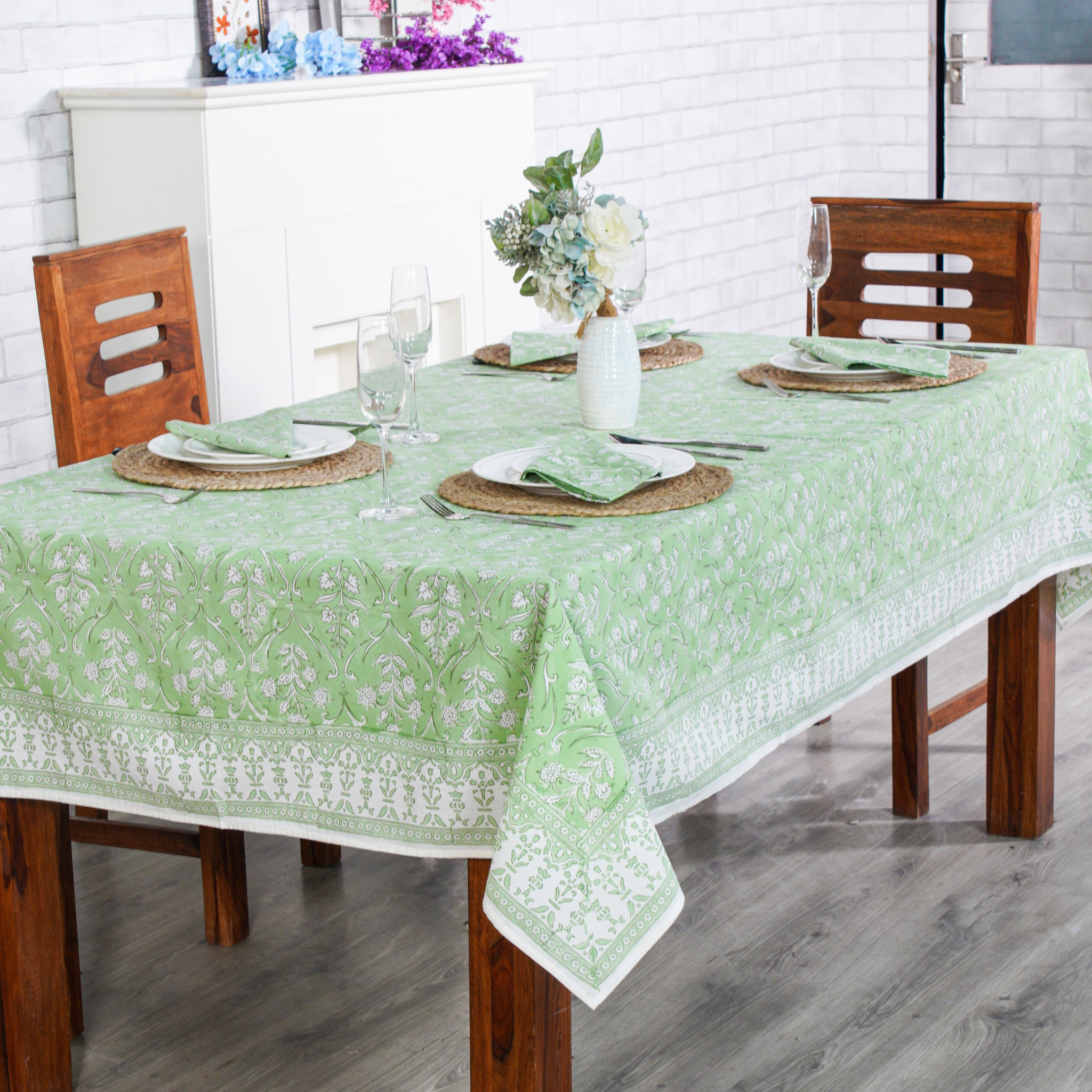 Dining table set with a green floral tablecloth, wooden chairs, and white plates.