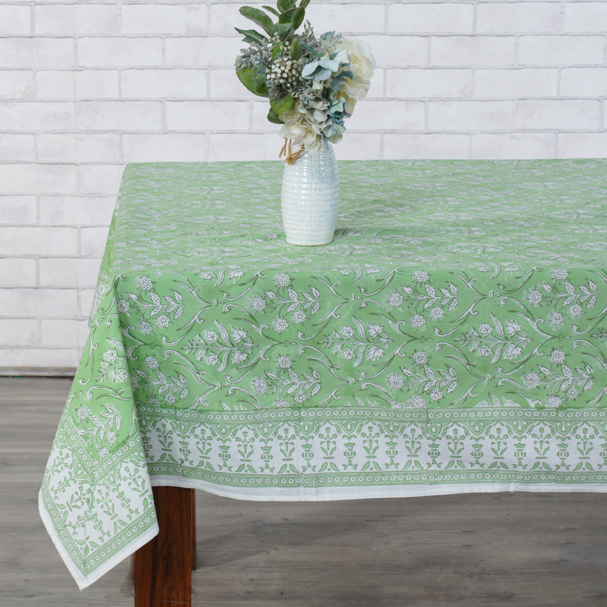 Green and white patterned tablecloth on a wooden table with a vase of flowers against a white brick wall.