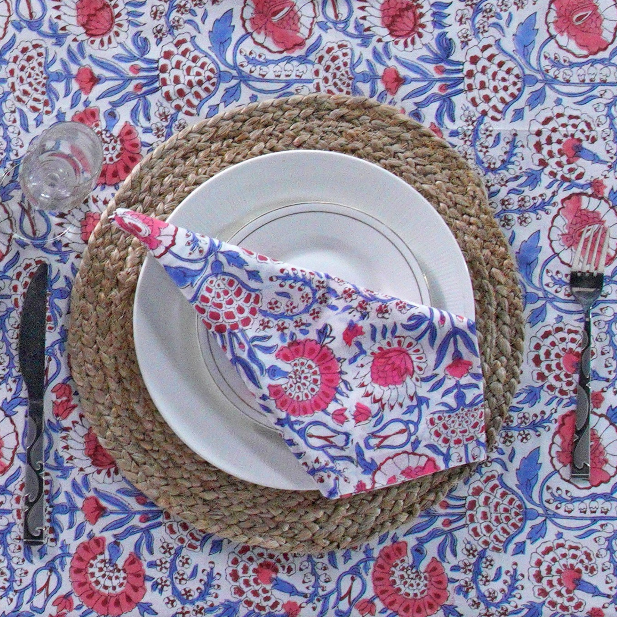 Table setting with a floral napkin, white plate, and cutlery on a patterned tablecloth.