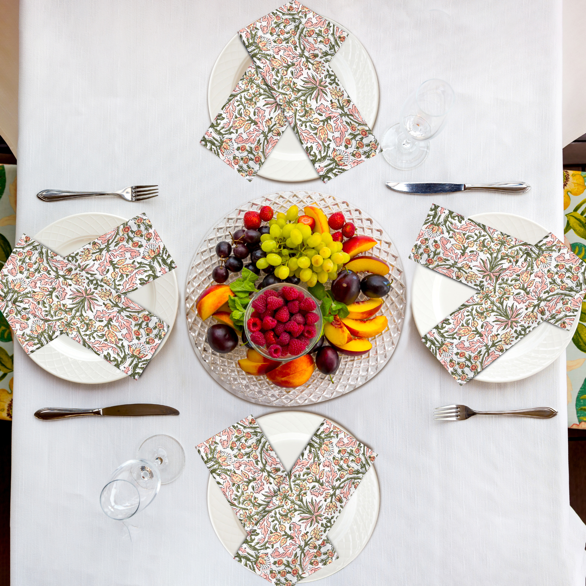 Table setting with floral plates, fruit salad, and cutlery on a white tablecloth.