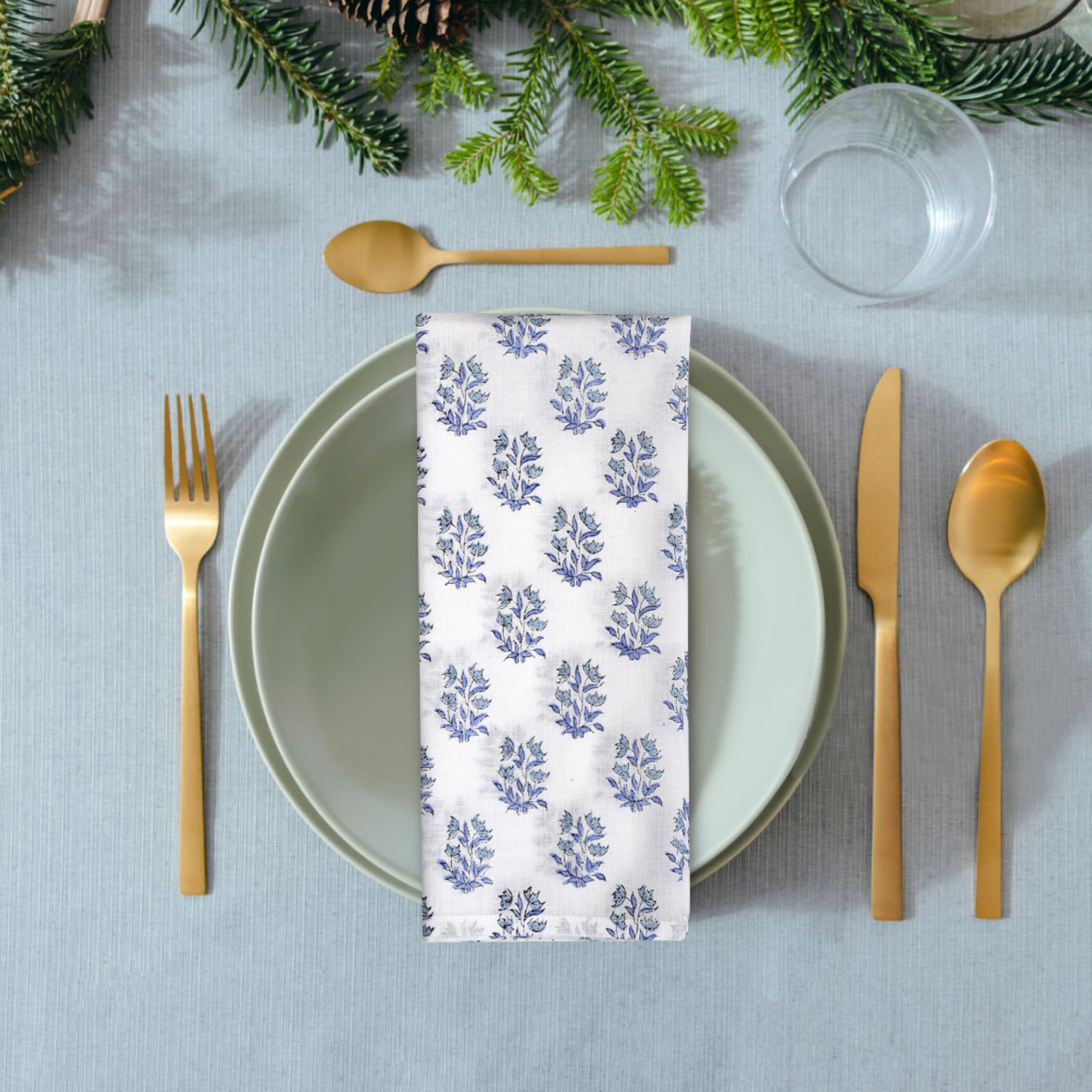 Table setting with a floral-patterned napkin, gold cutlery, and greenery on a gray tablecloth.