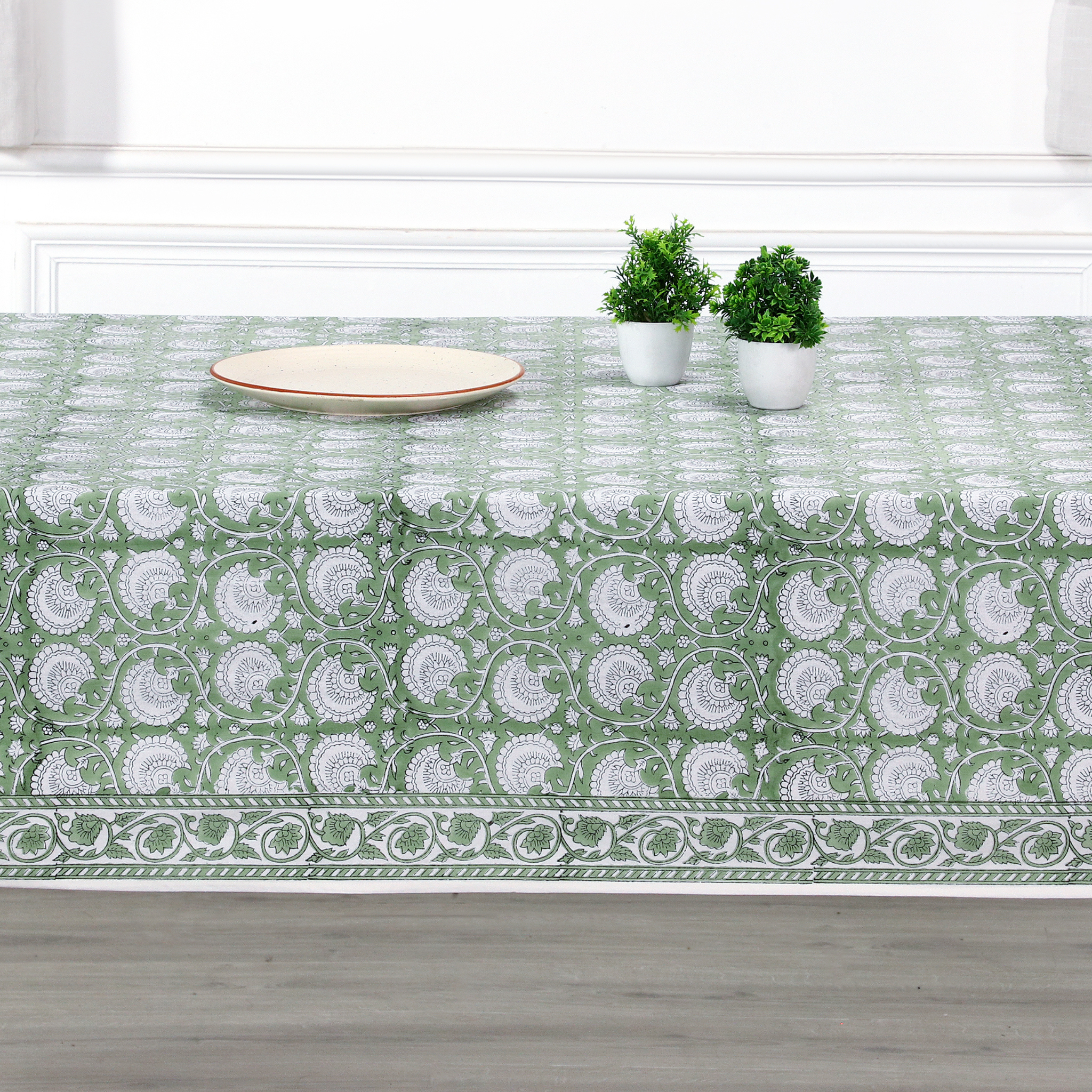 Green and white patterned tablecloth on a wooden table with plants and a plate.