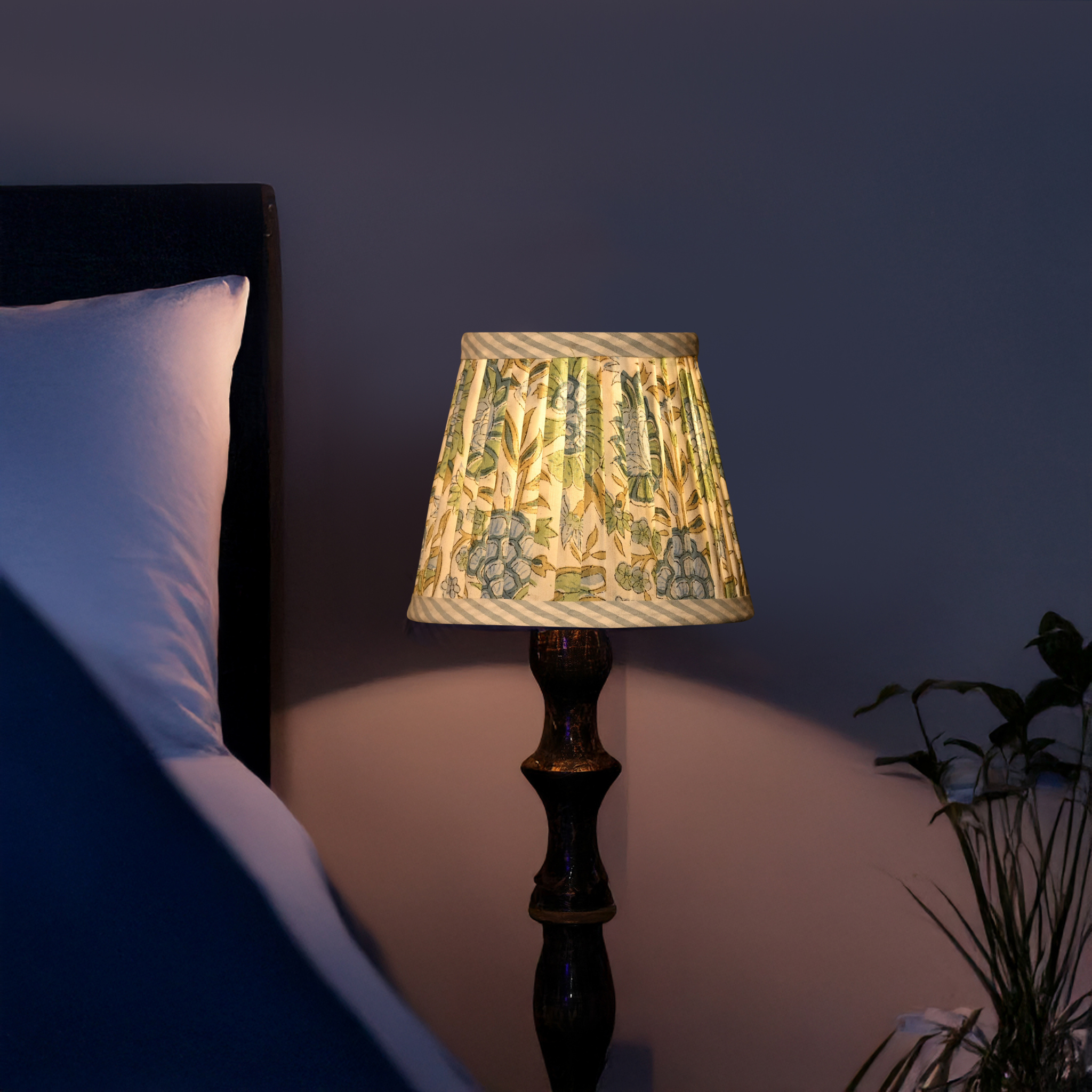 Lamp with patterned shade on a bedside table next to a bed in a dimly lit room.