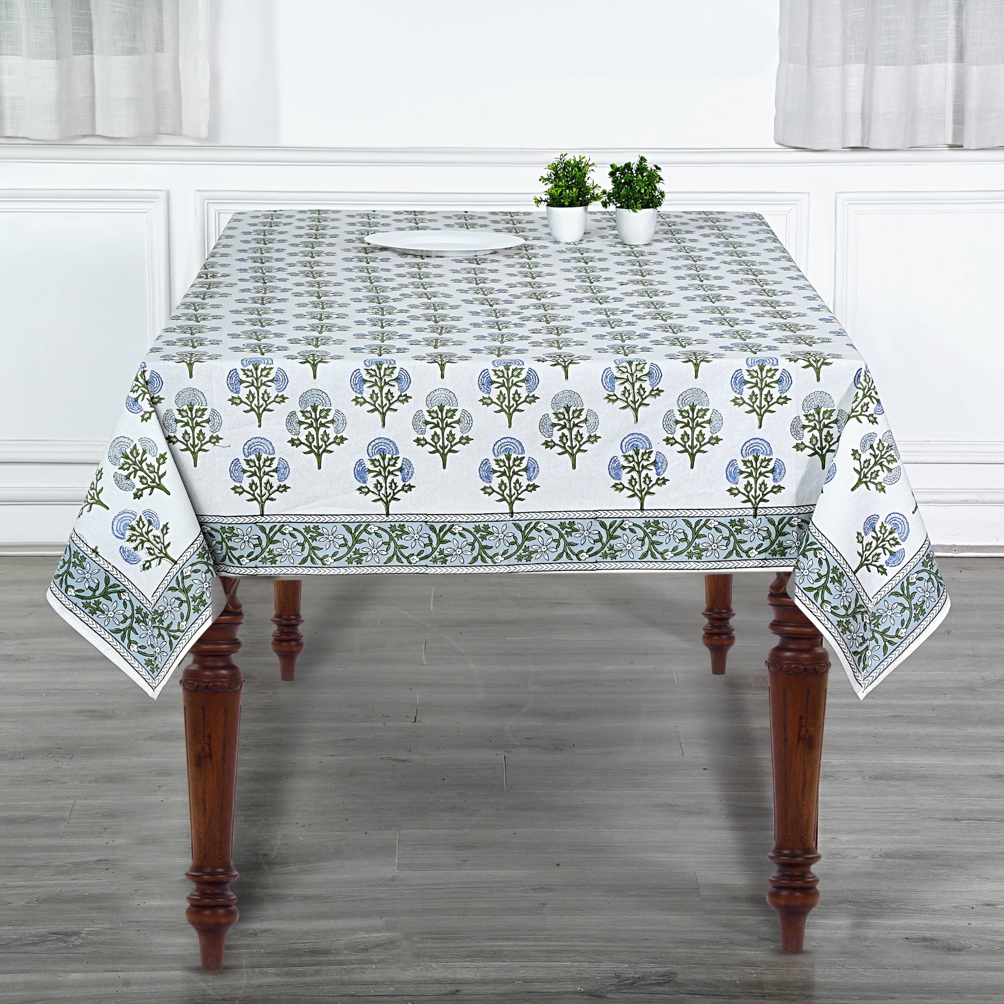 Tablecloth with floral pattern on a wooden table in a room with white walls and curtains.
