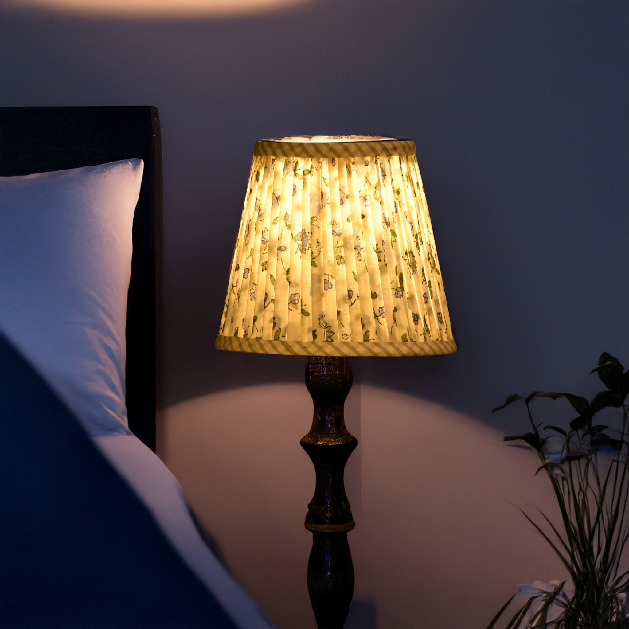 Lamp with a textured shade on a bedside table next to a bed in a dimly lit room.