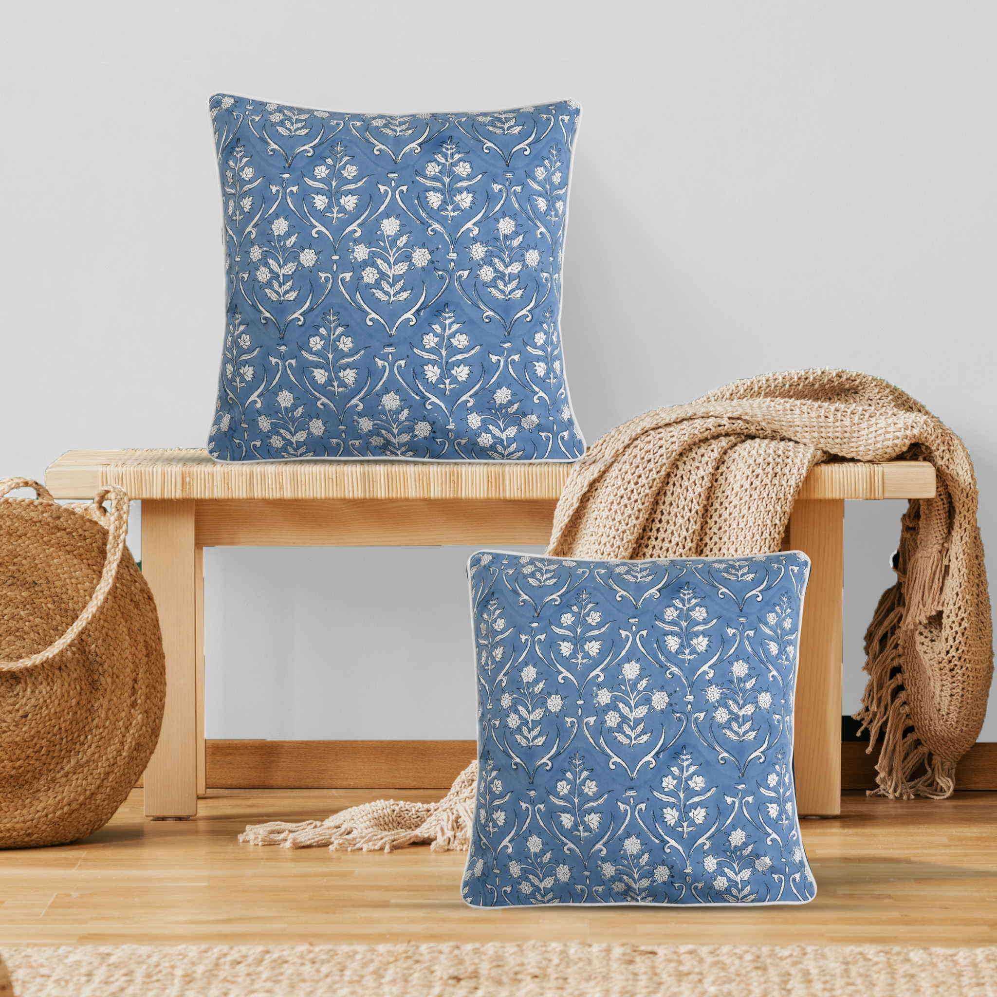 Two blue patterned pillows on a wooden stool with a beige woven basket and blanket.