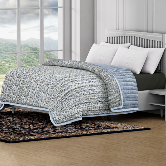 Bed with a textured quilt and white pillows in a room with large windows showing a scenic view.