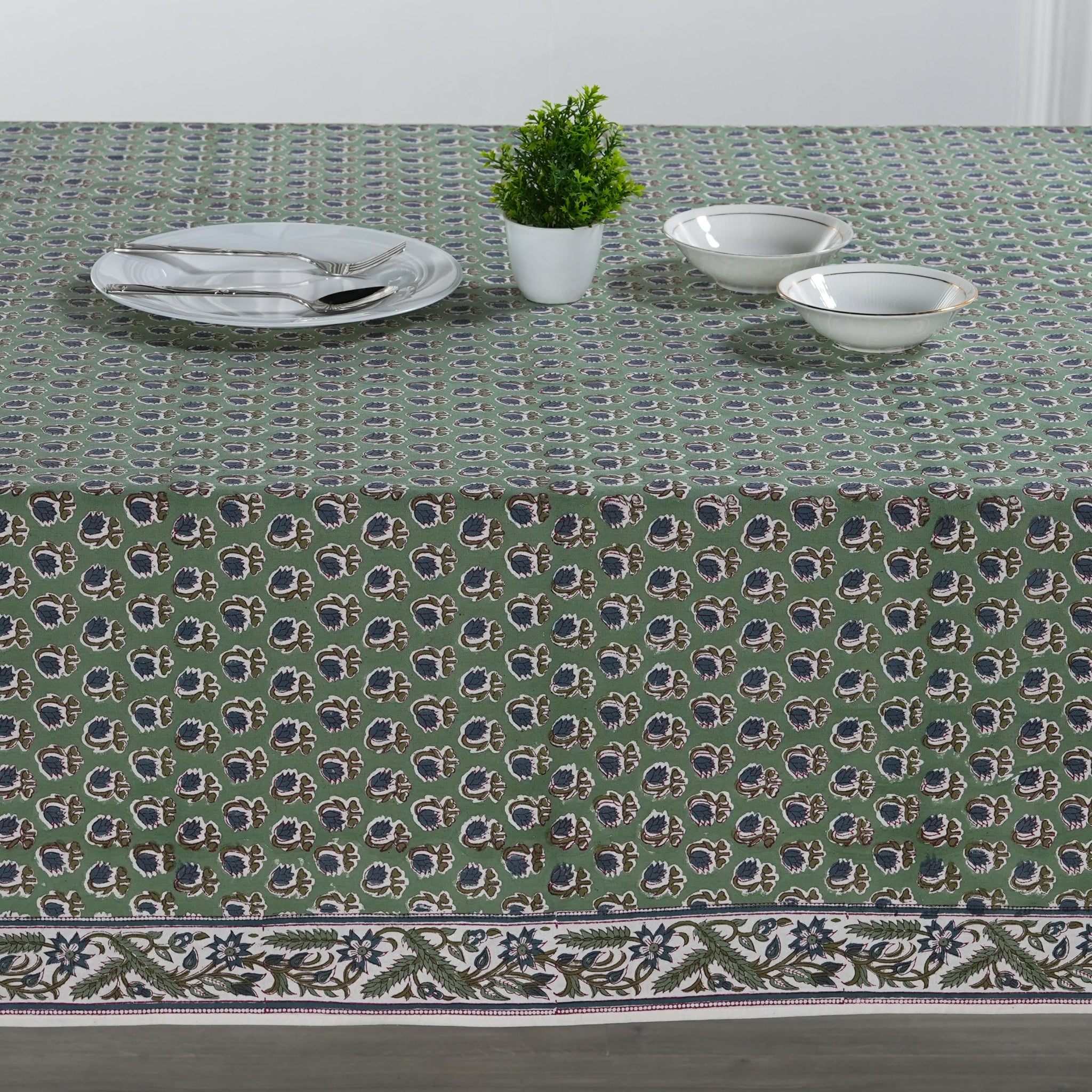 Green patterned tablecloth with white plates, silverware, and a small plant on a table.
