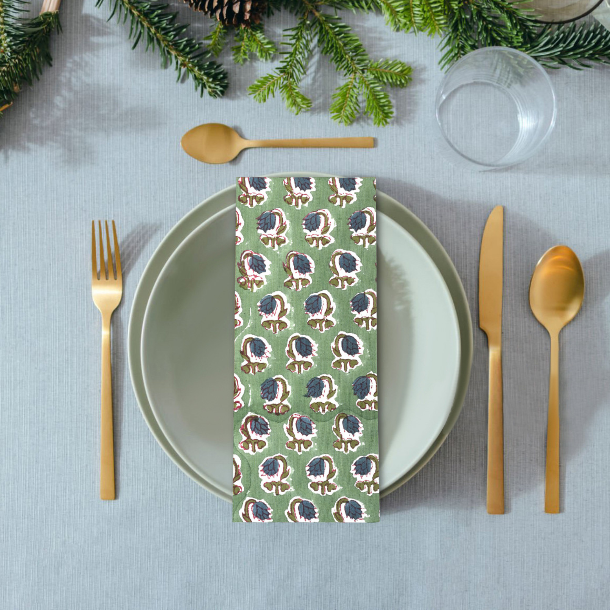 Table setting with a patterned napkin, gold cutlery, and greenery on a gray tablecloth.