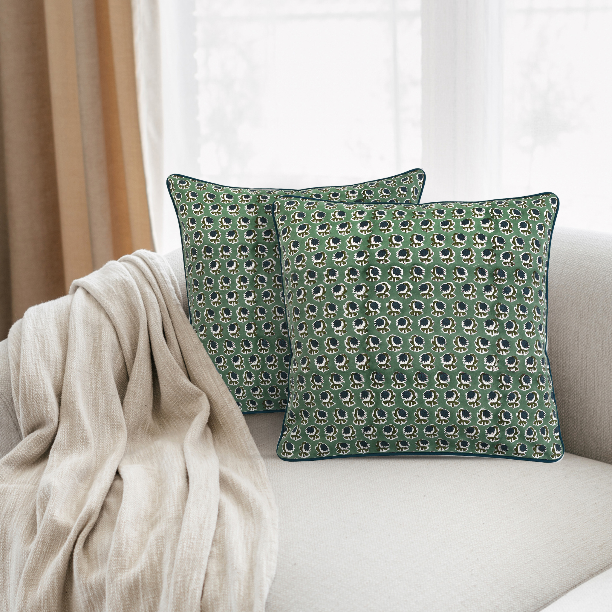 Two green patterned pillows on a beige sofa with a neutral curtain in the background