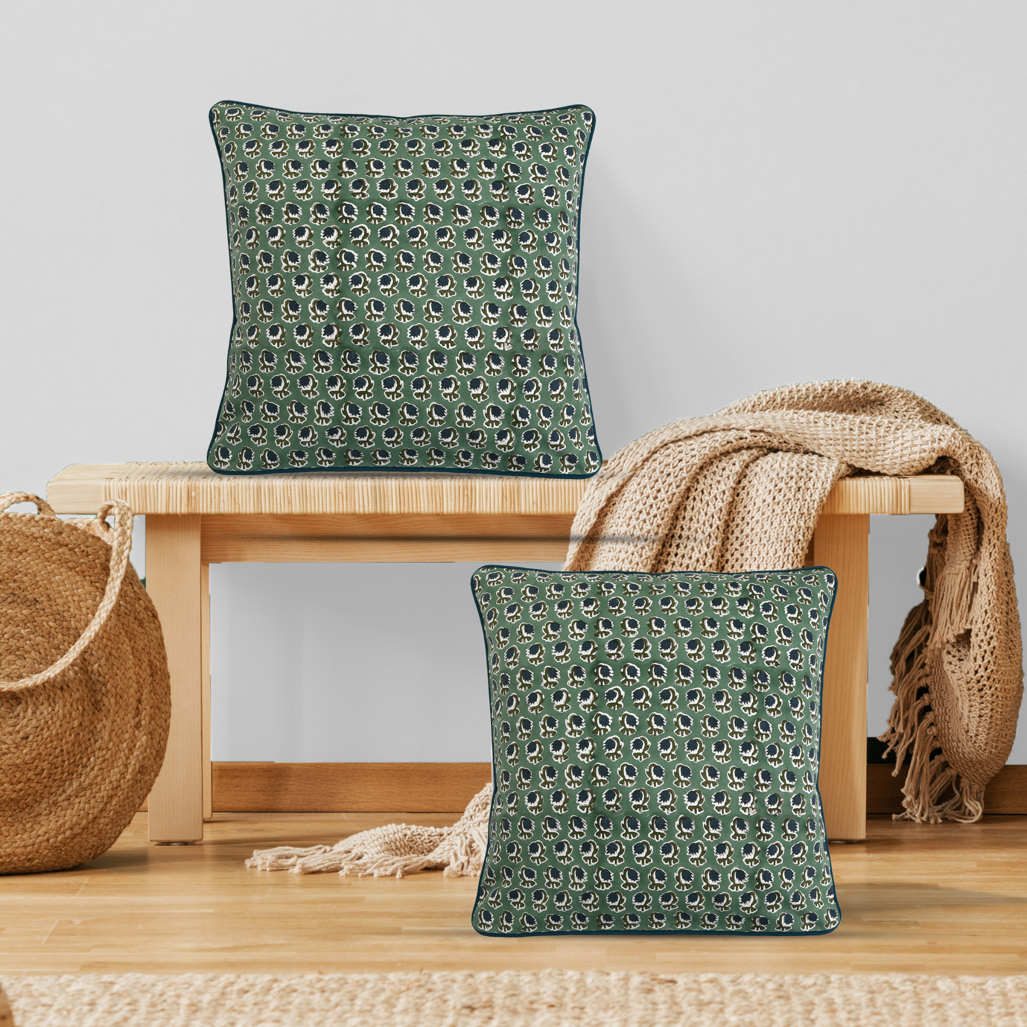 Two green patterned cushions on a wooden stool with a beige blanket and woven basket.