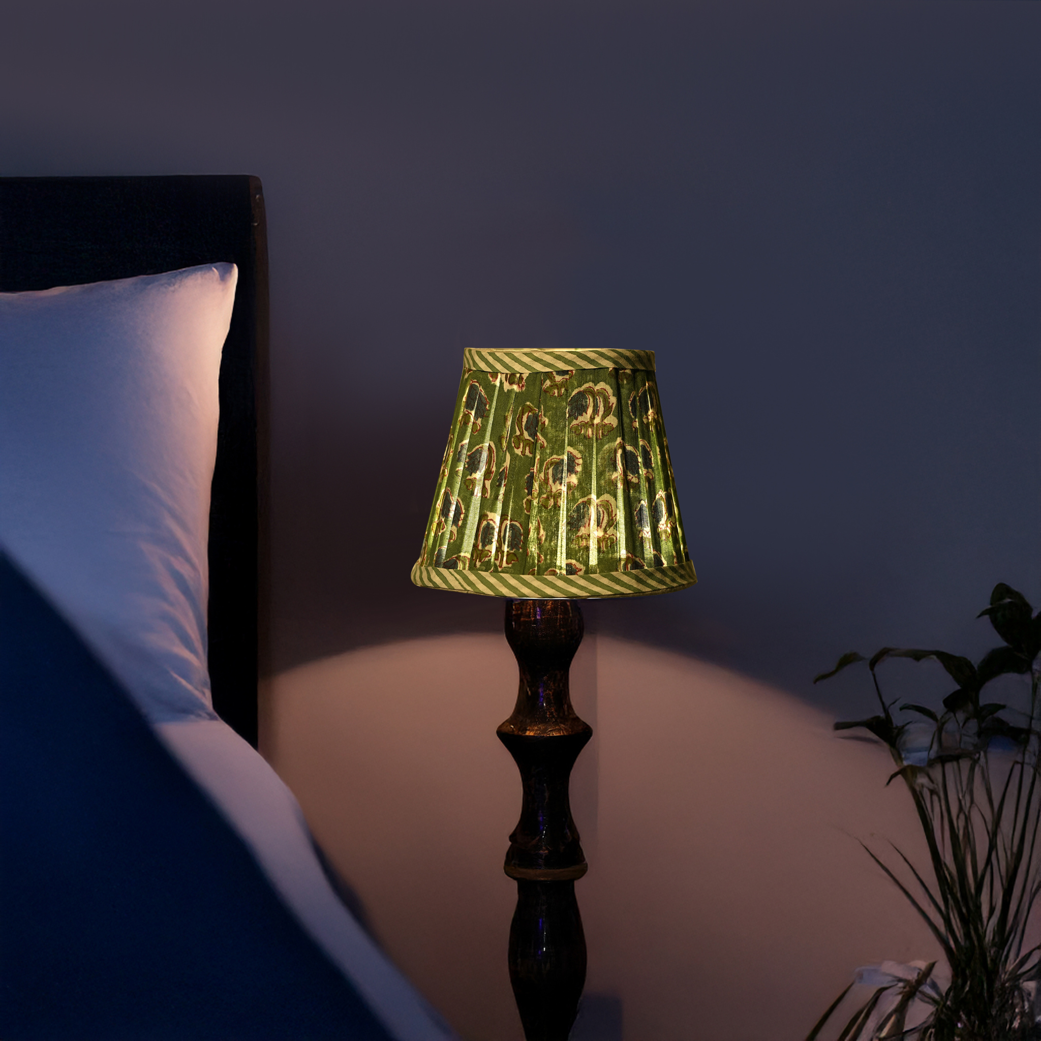 Lamp with a green patterned shade on a bedside table next to a bed in a dimly lit room.