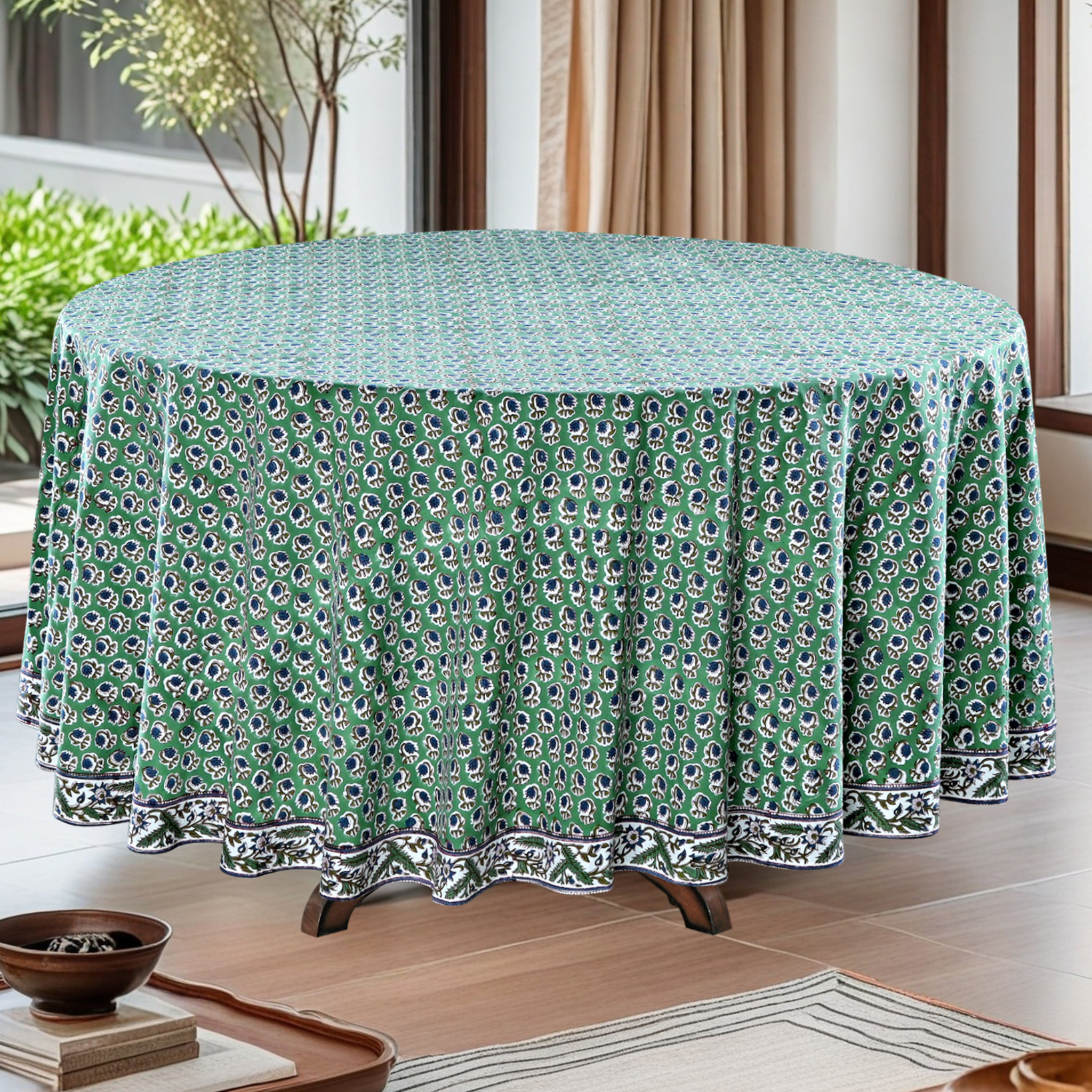 Round table with a green patterned tablecloth in a room with plants and curtains.