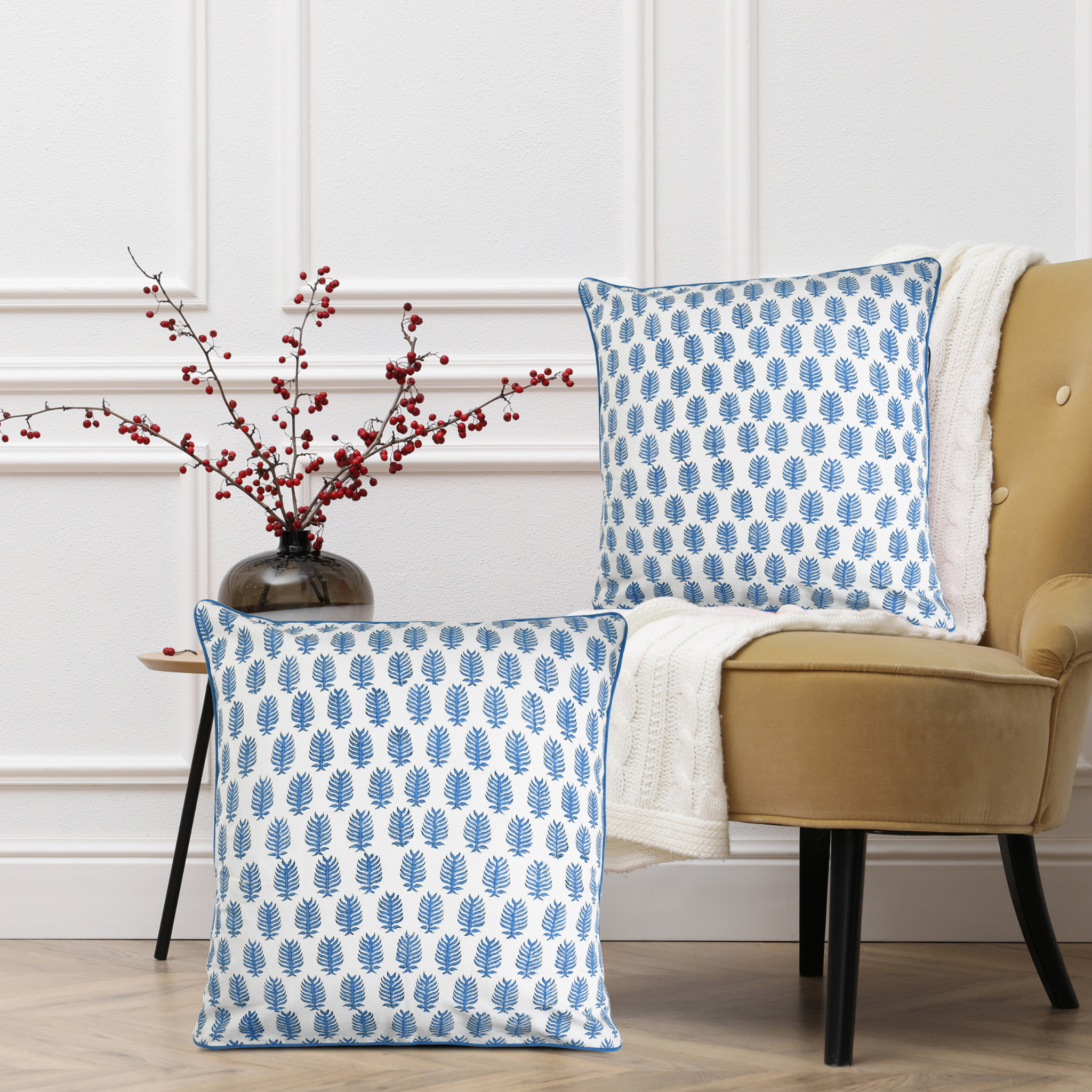 Two blue and white patterned pillows on a beige armchair with a vase of red berries on a side table.
