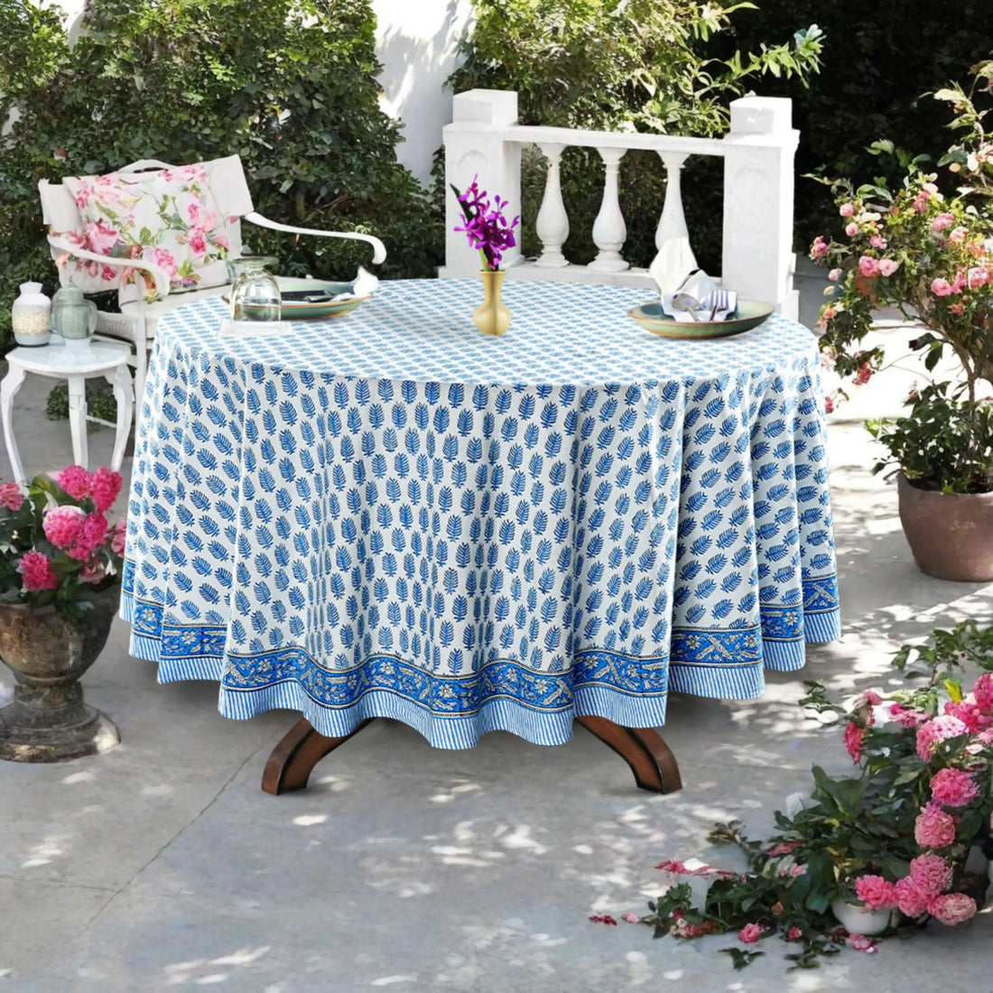 Outdoor setting with a table covered by a blue patterned tablecloth, surrounded by potted plants and chairs.