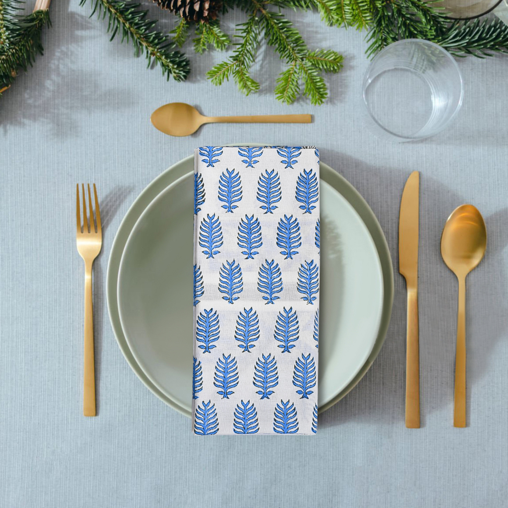 Table setting with a patterned blue and white napkin, gold cutlery, and greenery on a gray tablecloth.