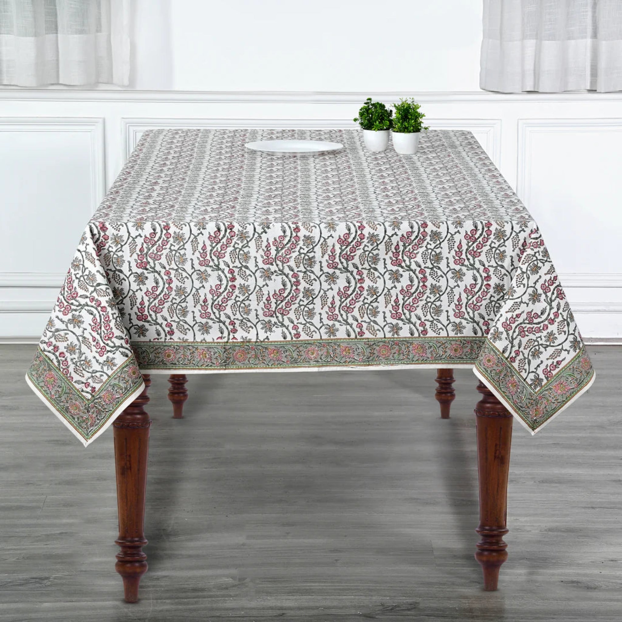 Tablecloth with floral pattern on a wooden table in a room with white walls and curtains.