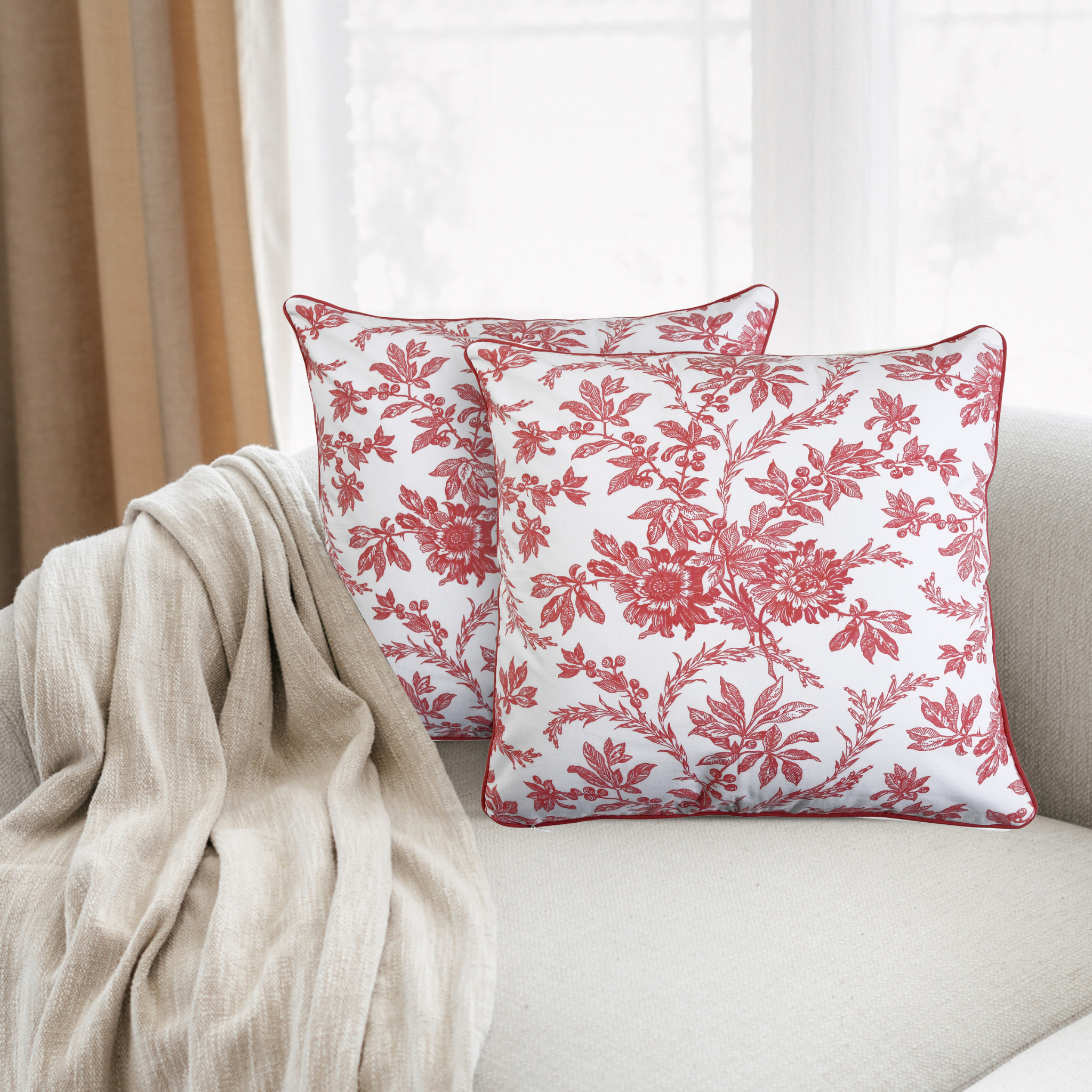Two red floral patterned pillows on a beige couch with a neutral curtain in the background.