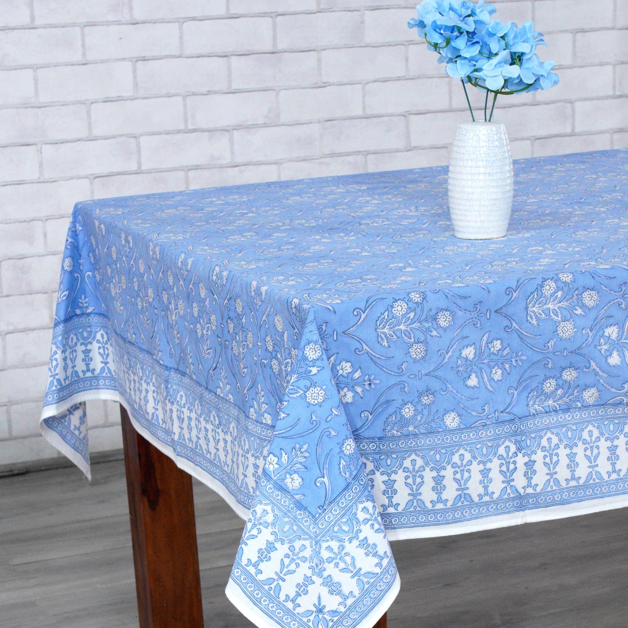 Blue patterned tablecloth on a wooden table with a white vase containing blue flowers against a white brick wall.