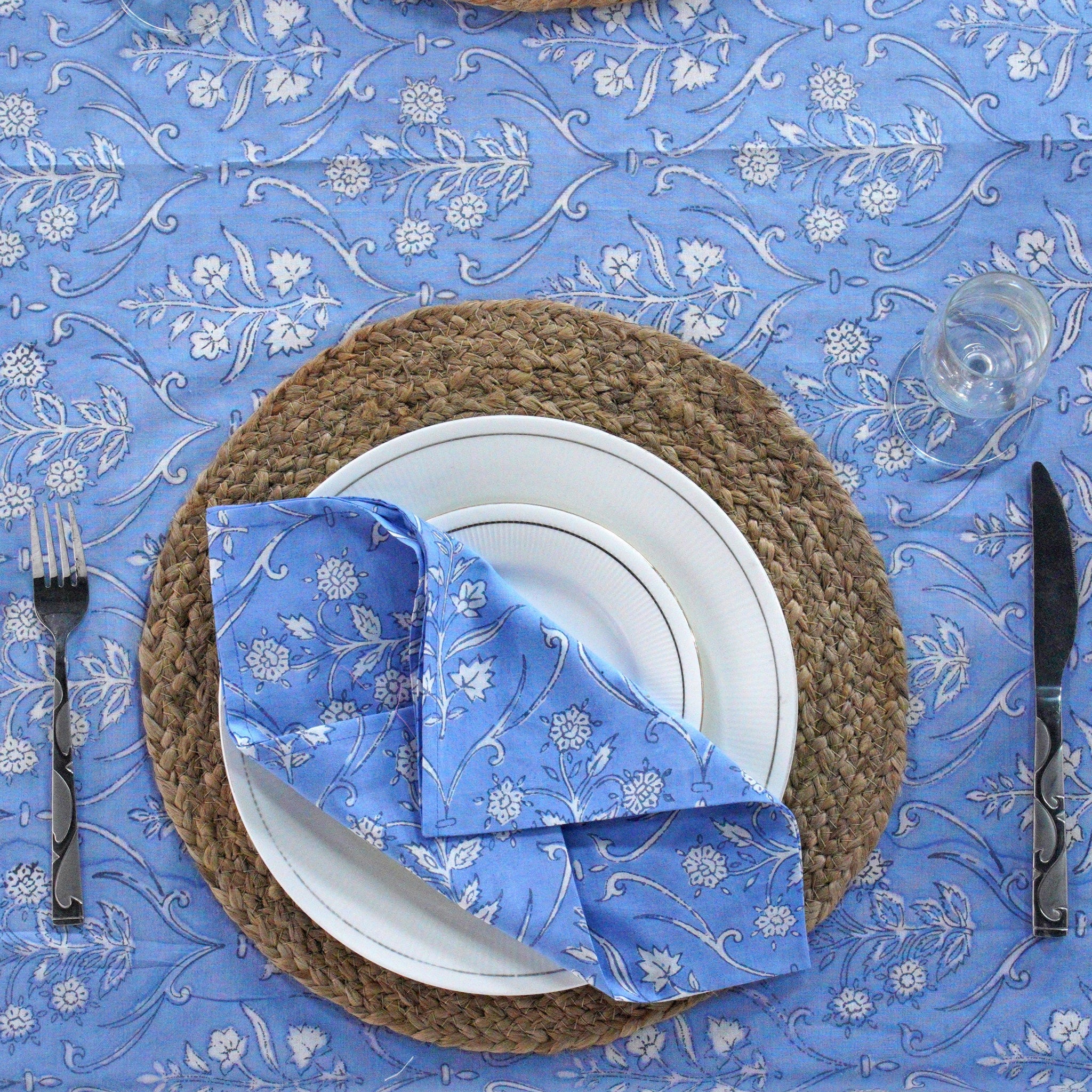 Table setting with a blue floral tablecloth, white plate, woven placemat, and blue napkin.