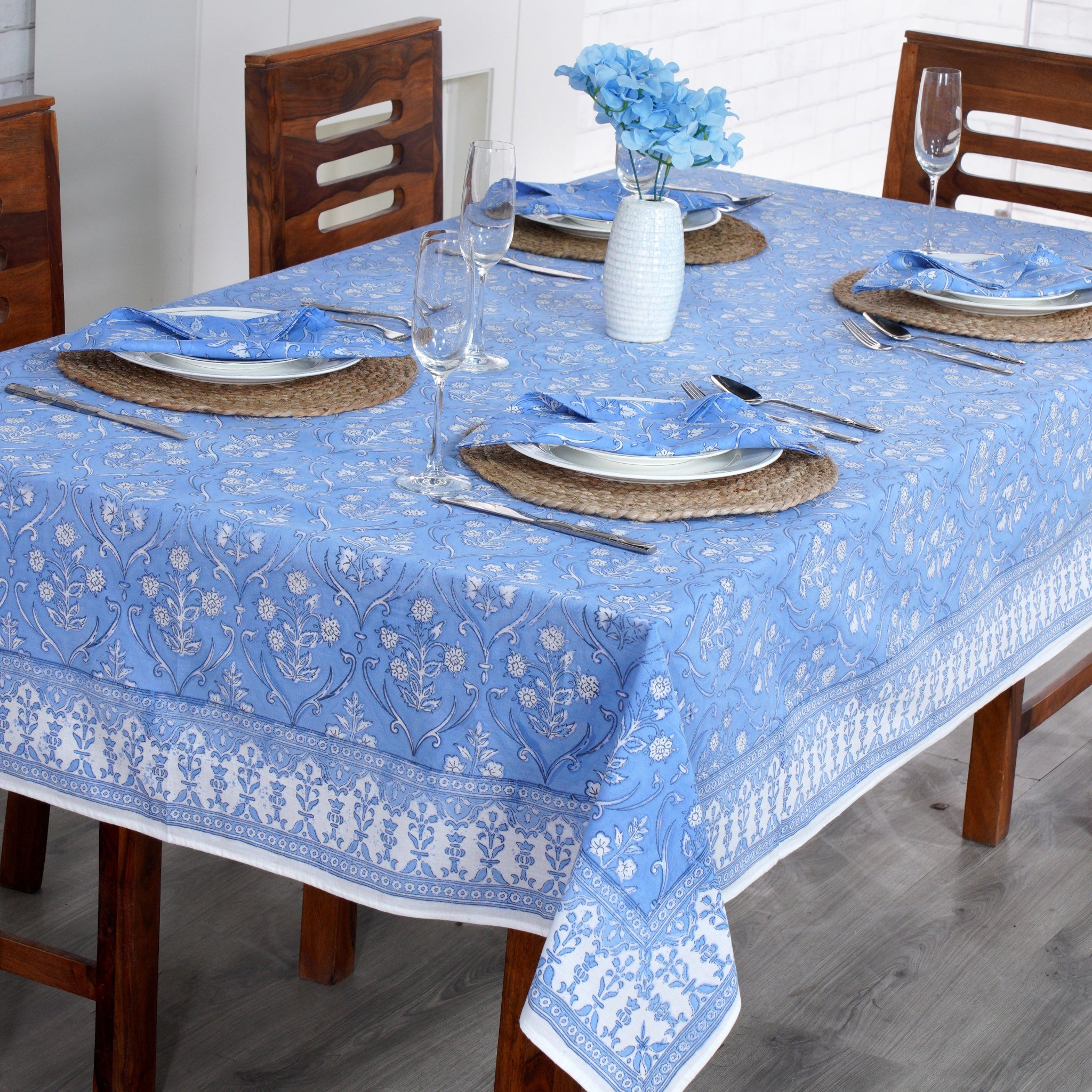 Dining table set with a blue floral tablecloth, wooden chairs, and white plates.