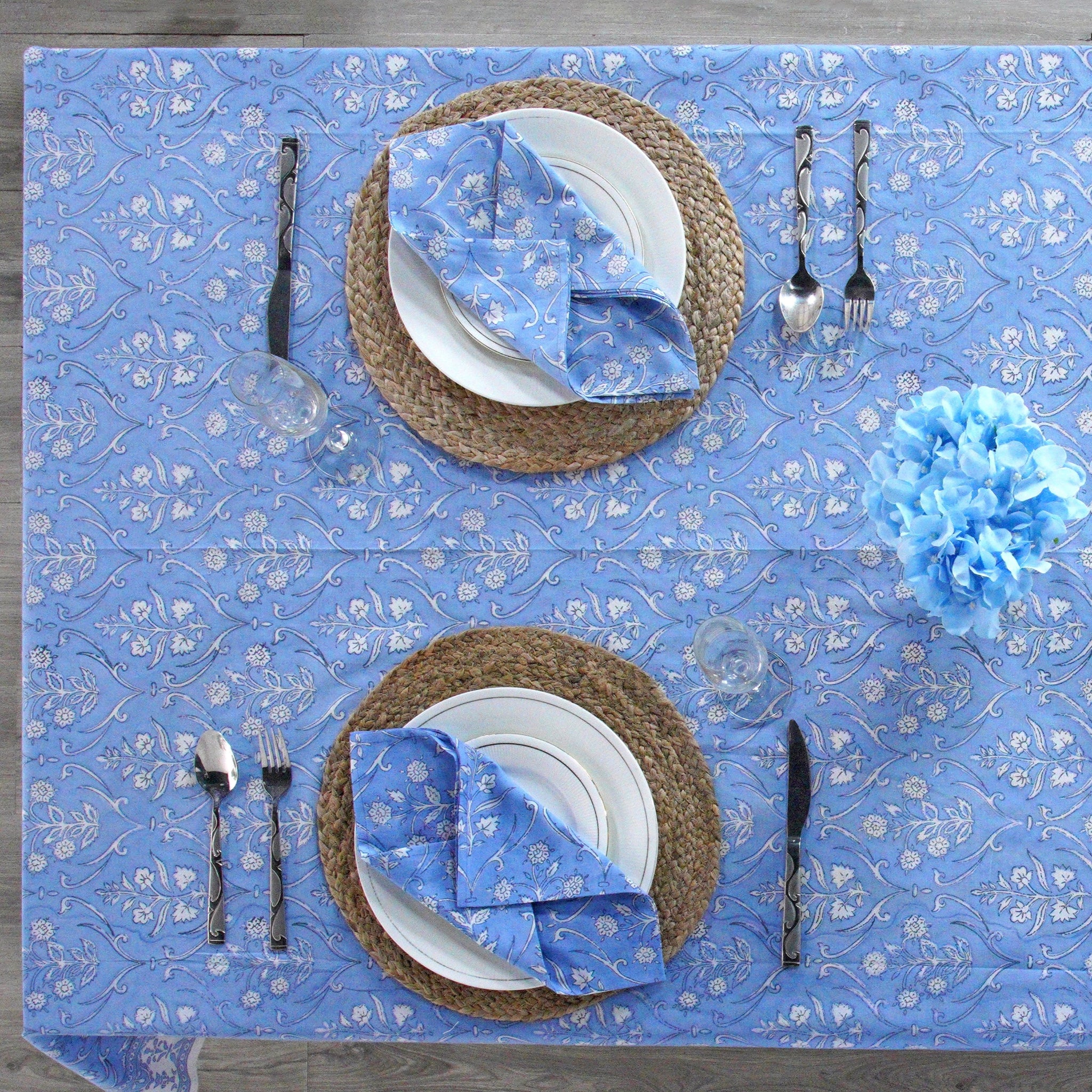 Set table with blue placemats, white plates, and silverware on a blue floral patterned tablecloth.