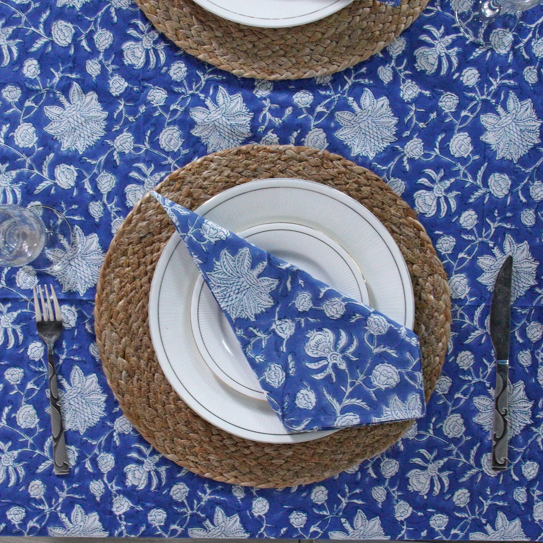 Table setting with blue floral tablecloth, woven placemat, white plates, and silverware.