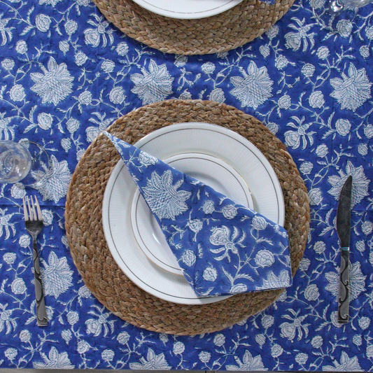 Table setting with blue floral tablecloth, woven placemat, white plates, and silverware.