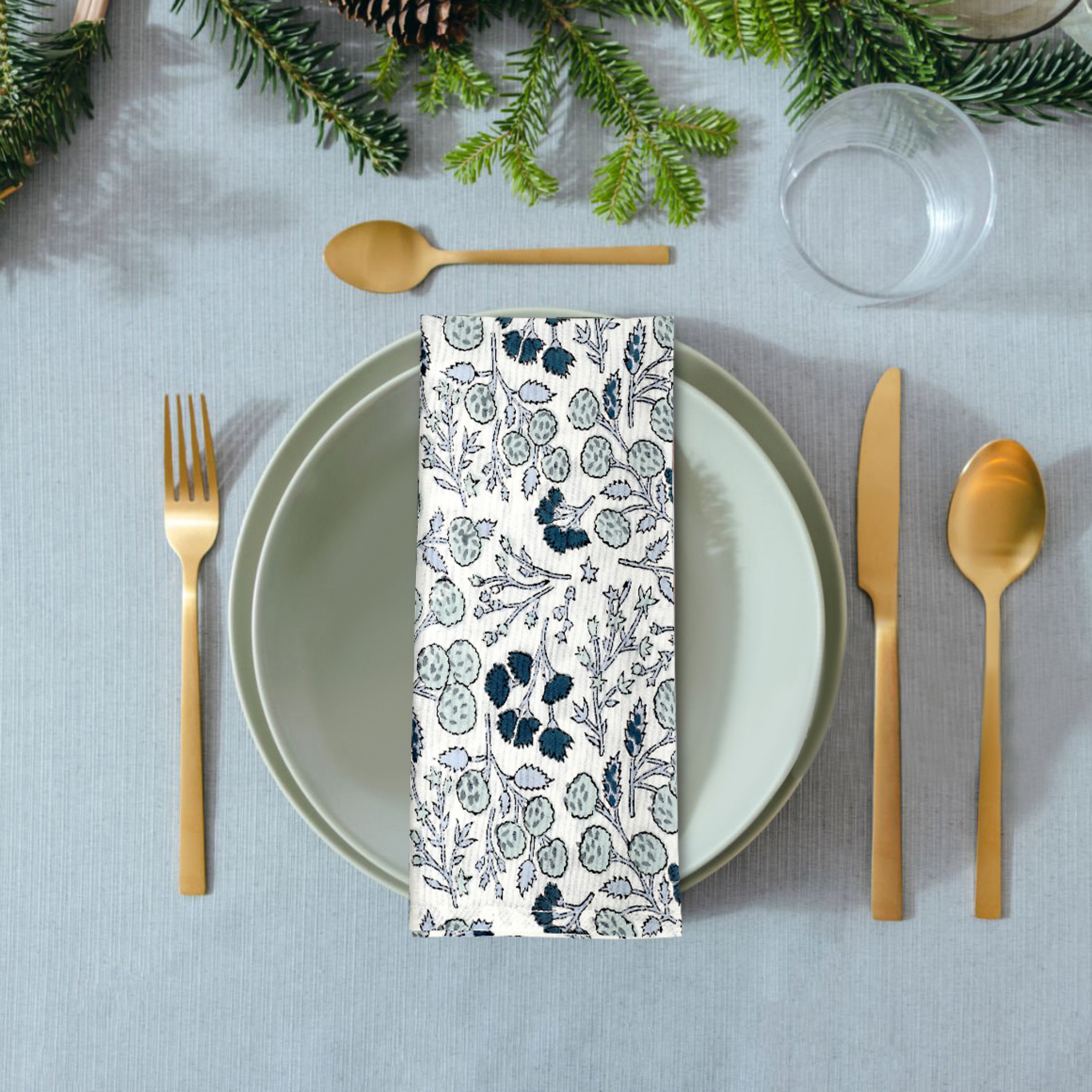 Table setting with a floral-patterned napkin, gold cutlery, and greenery on a gray tablecloth.