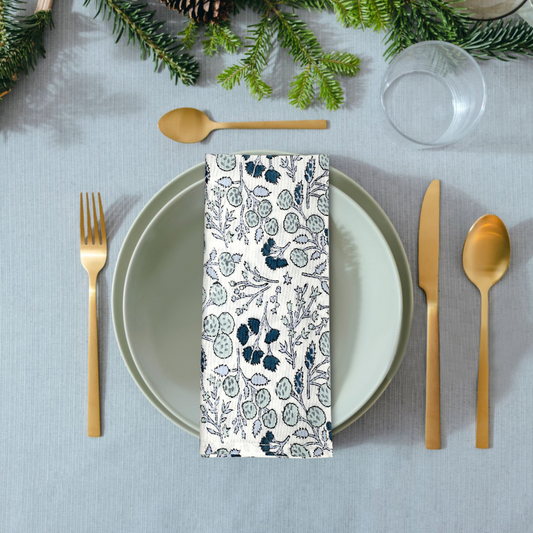 Table setting with a floral-patterned napkin, gold cutlery, and greenery on a gray tablecloth.