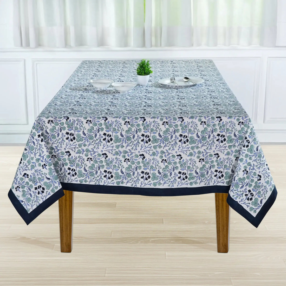 Tablecloth with floral pattern on a wooden table in a room with white walls and curtains.