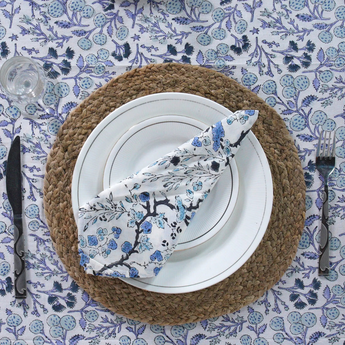 Table setting with a woven placemat, white plates, and a floral-patterned napkin on a blue and white tablecloth.