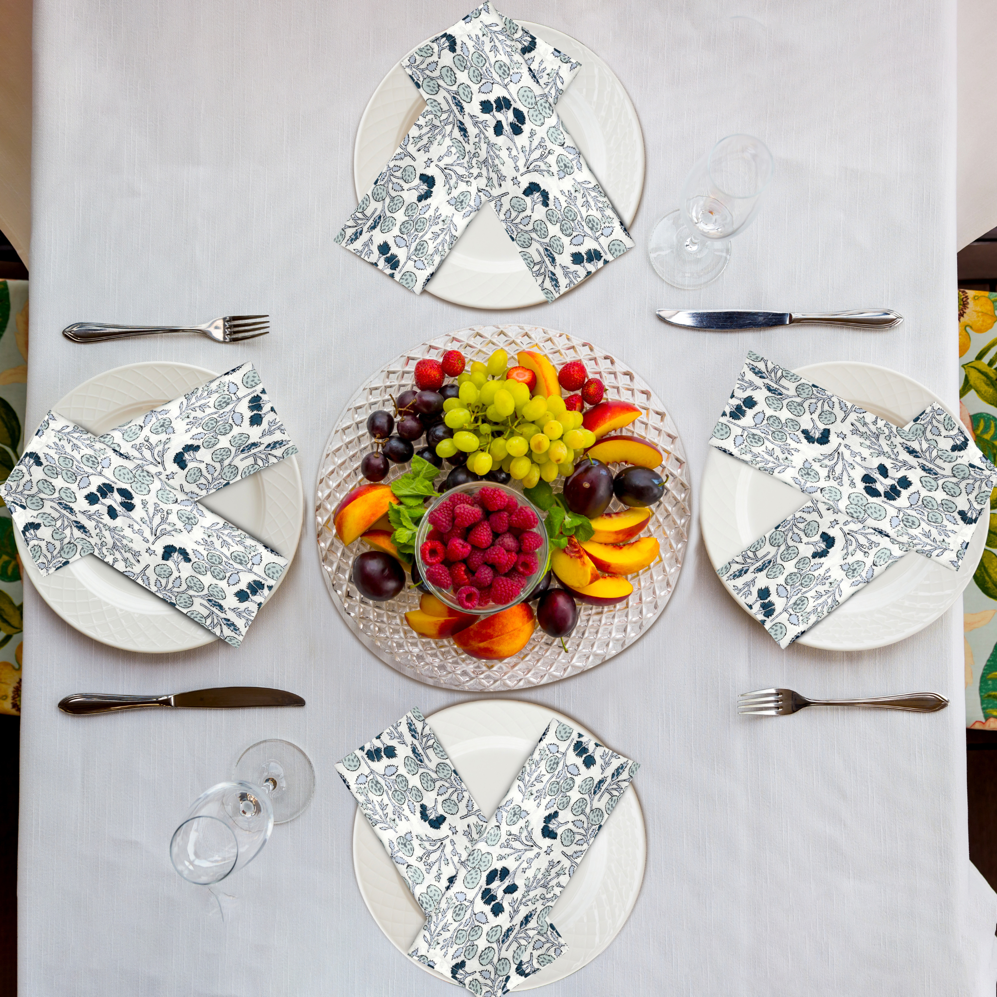 Table setting with floral napkins, fruit platter, and cutlery on a white tablecloth.
