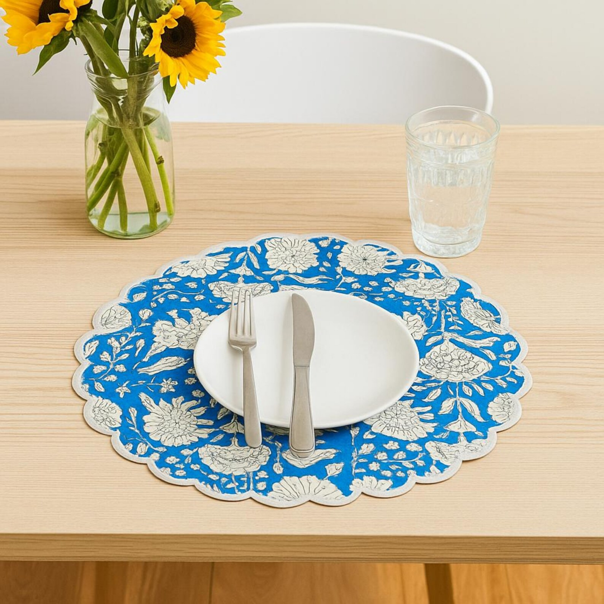 Blue and white floral-patterned placemat on a wooden table with a glass and vase of sunflowers.