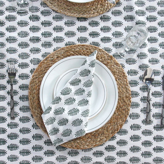 Table setting with woven placemat, white plates, and silverware on a leaf-patterned tablecloth.